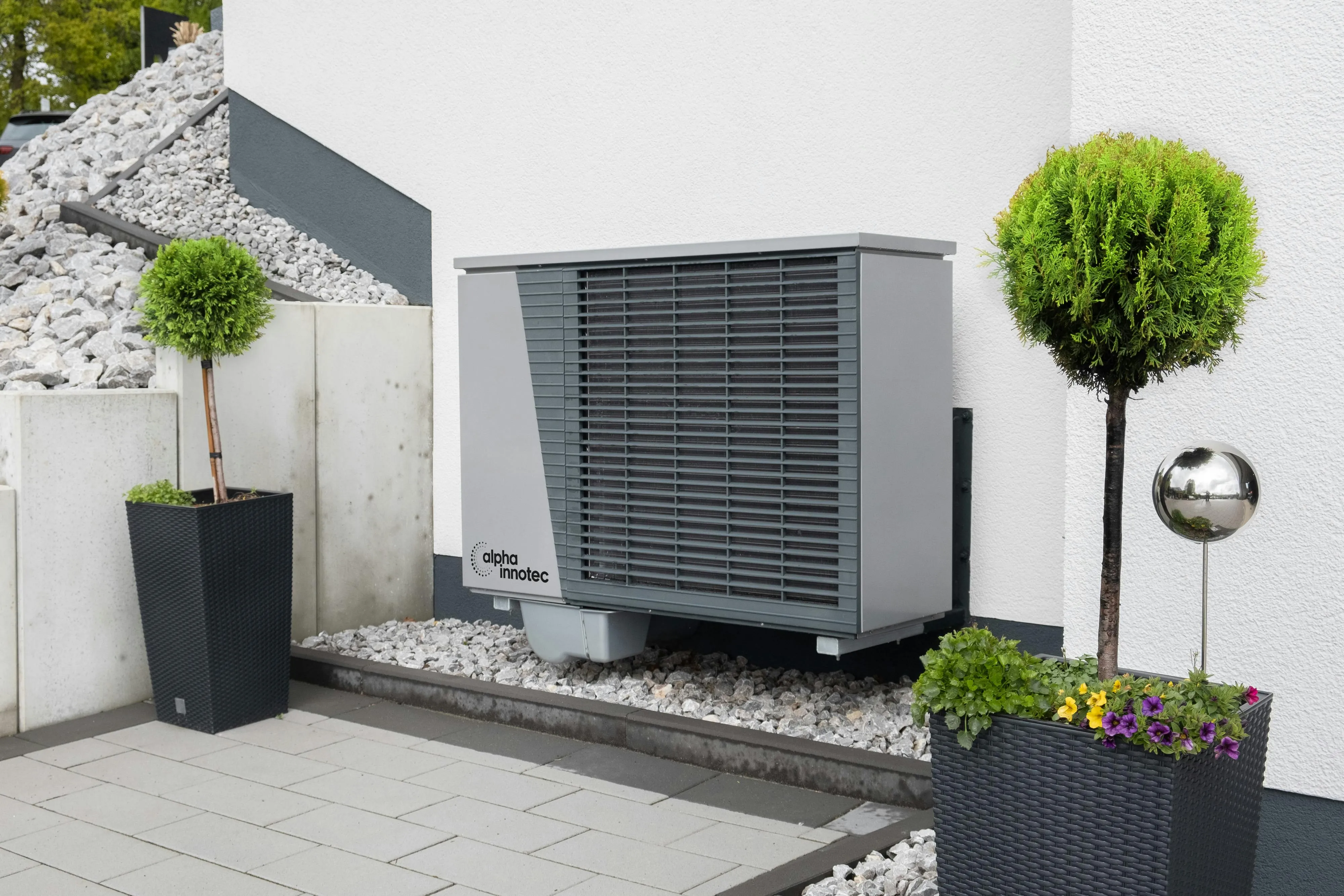 Gray alpha innotec heat pump unit mounted on an exterior white wall between two potted topiary trees with flowers.