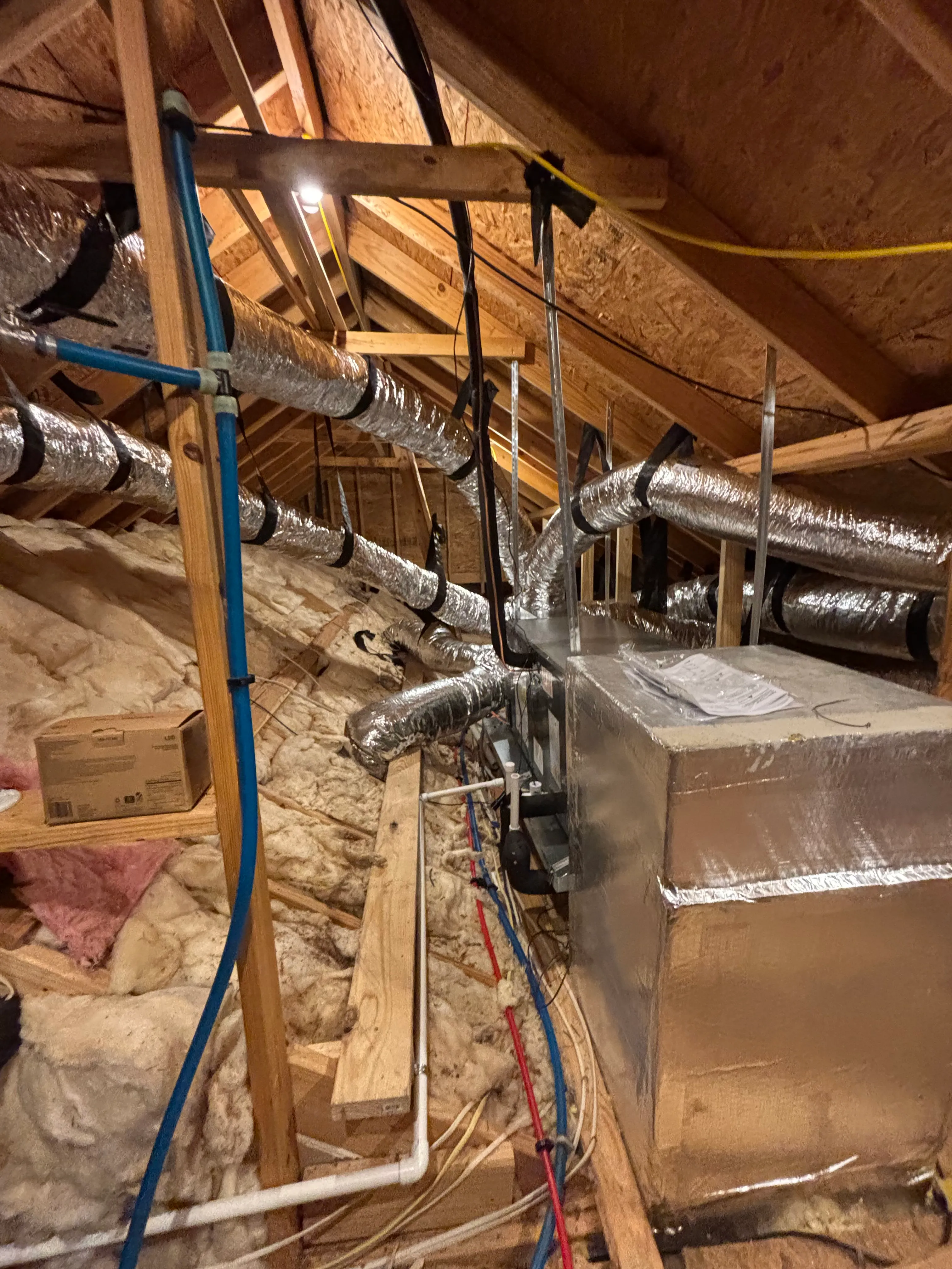Attic space with exposed wooden beams, insulation, and silver HVAC ductwork connected to a large metal unit.
