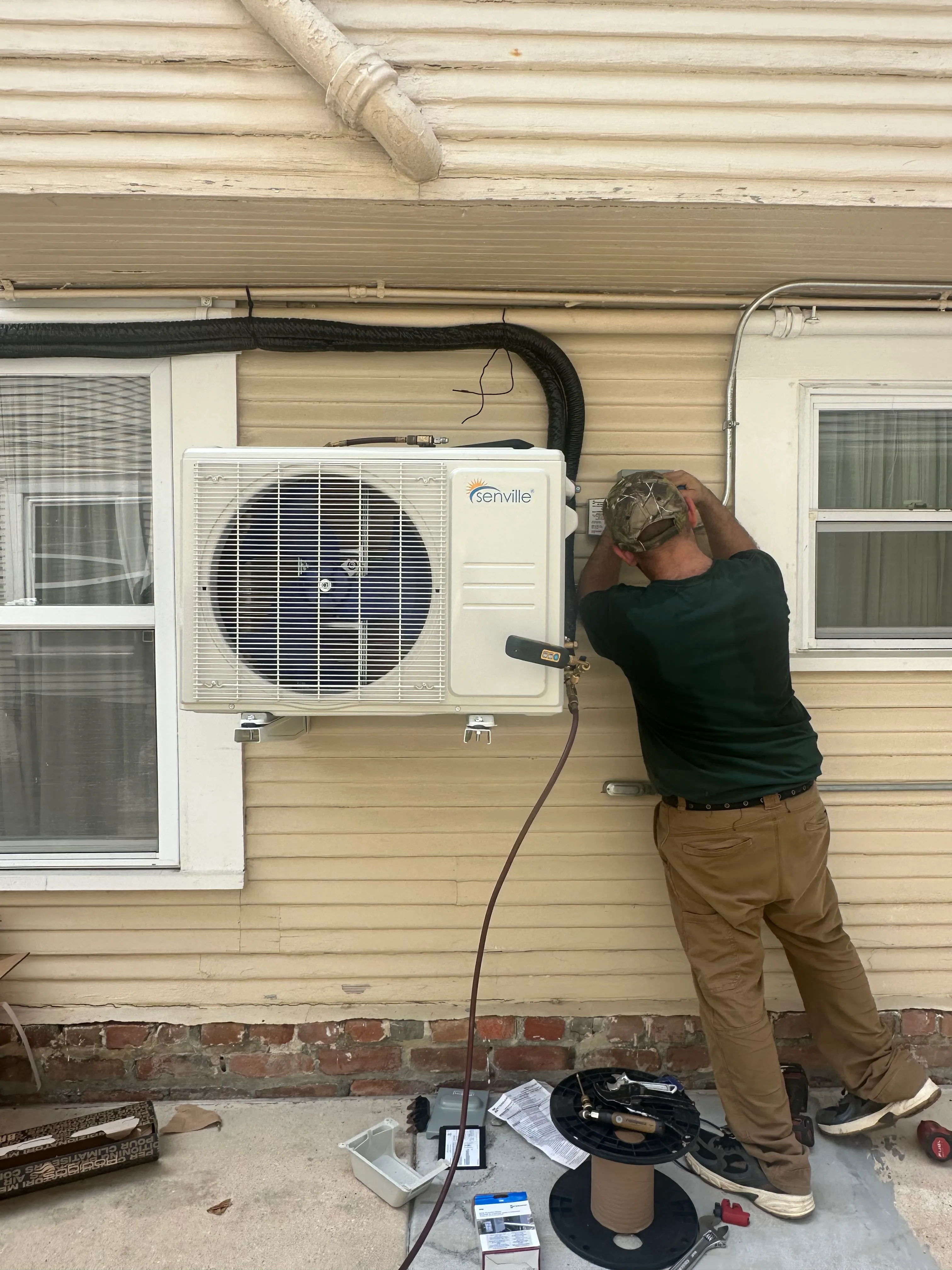Technician in a camouflage cap and brown pants installing a Senville air conditioning unit on the exterior wall of a beige house.