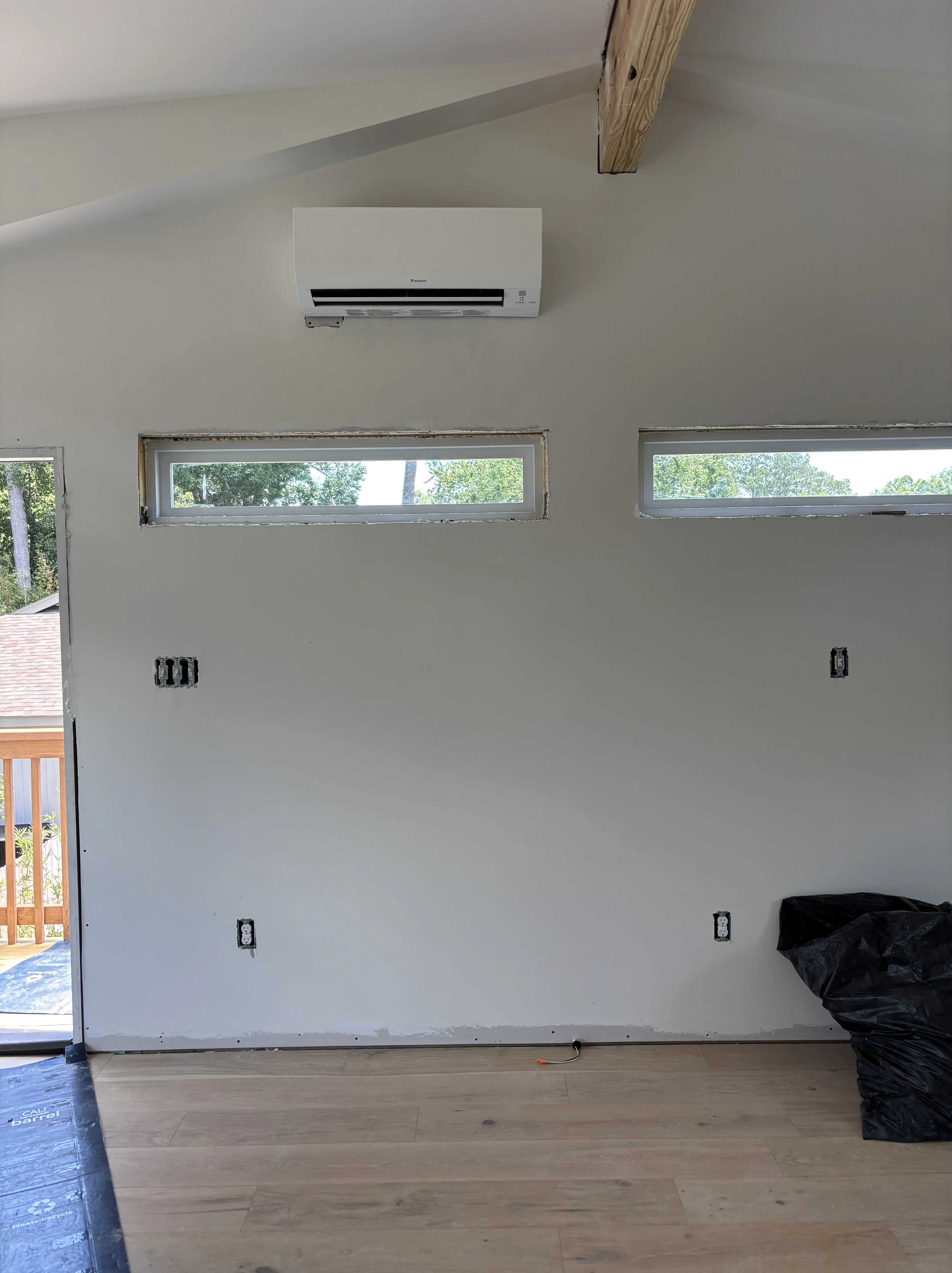 Interior wall with mounted air conditioning unit, two narrow horizontal windows, and unfinished electrical outlets in a room with wood flooring.