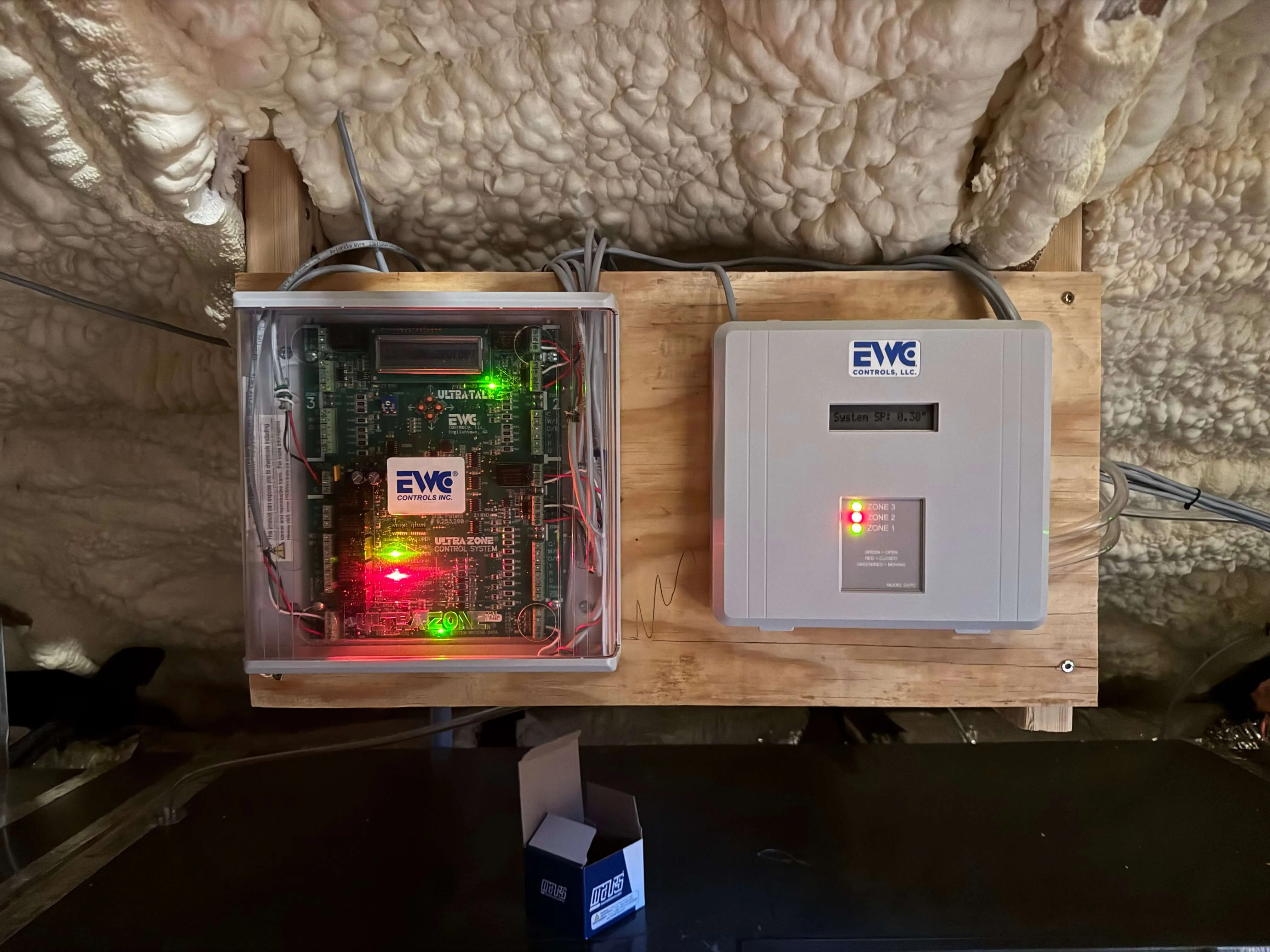 Two EWC control system units mounted on a wooden board against insulated wall, with lit indicator lights and connected wiring.