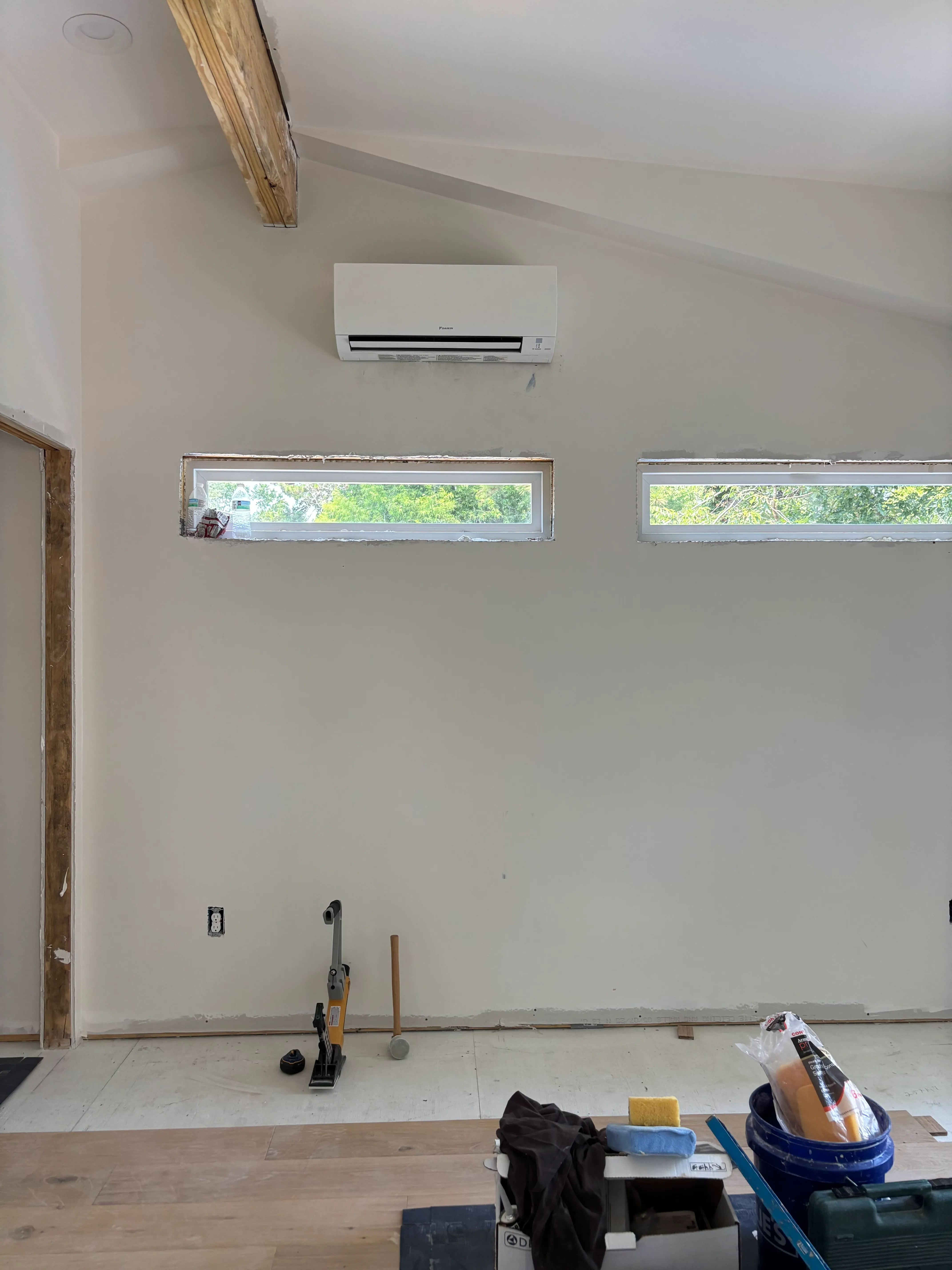 Interior wall with a mounted white air conditioning unit above two horizontal windows, with construction tools and materials on the floor in front.