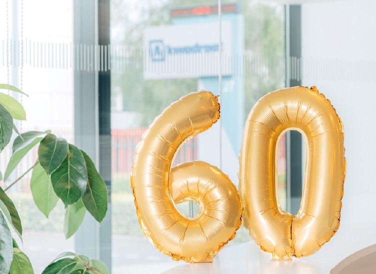 Gold foil balloons forming the number 60 are displayed next to a green plant, against a backdrop of large windows, suggesting a festive atmosphere.