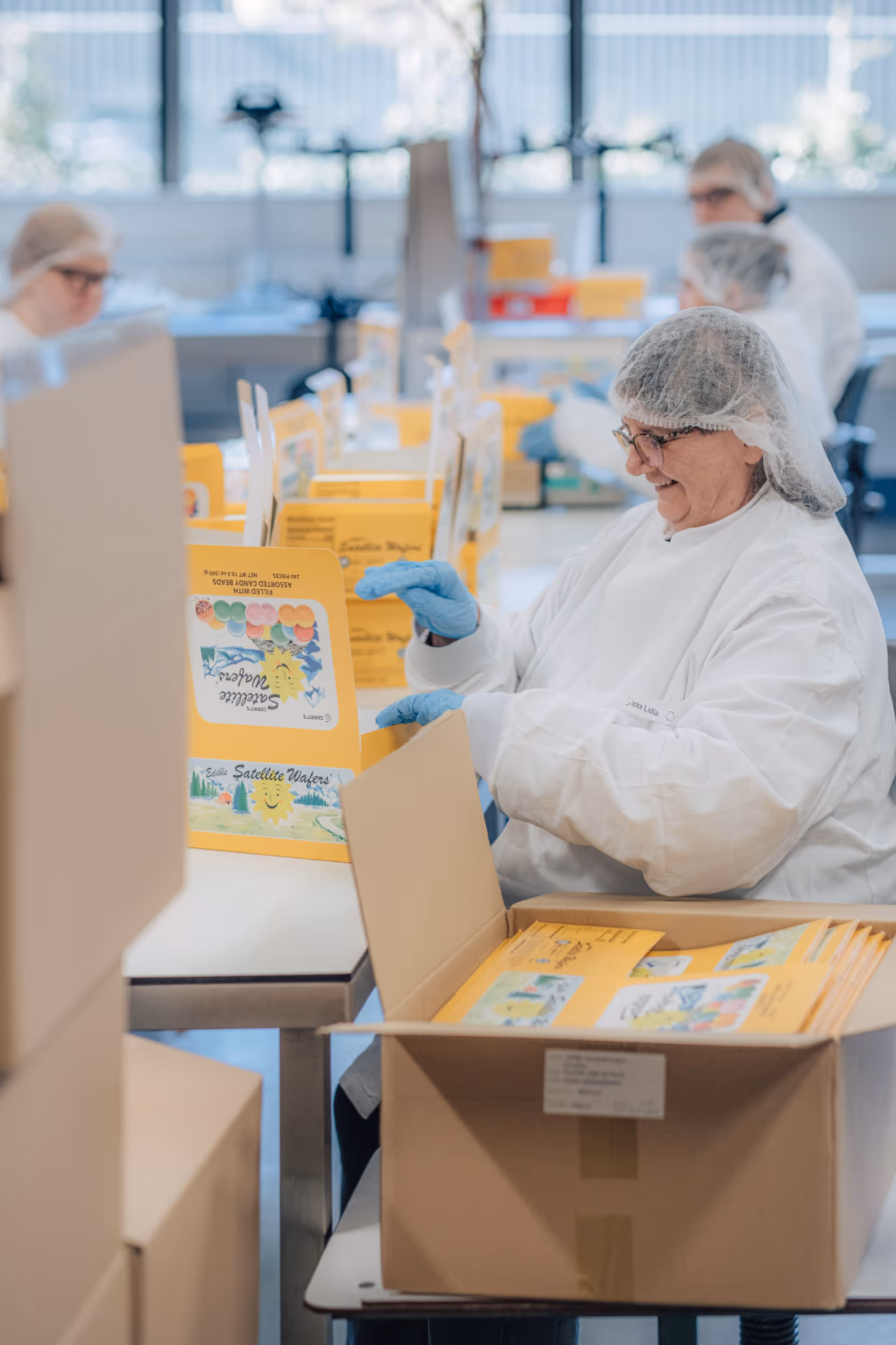 A person wearing protective clothing and a hairnet smiles as she assembles colorful packaging at a table. The atmosphere is focused and cheerful.