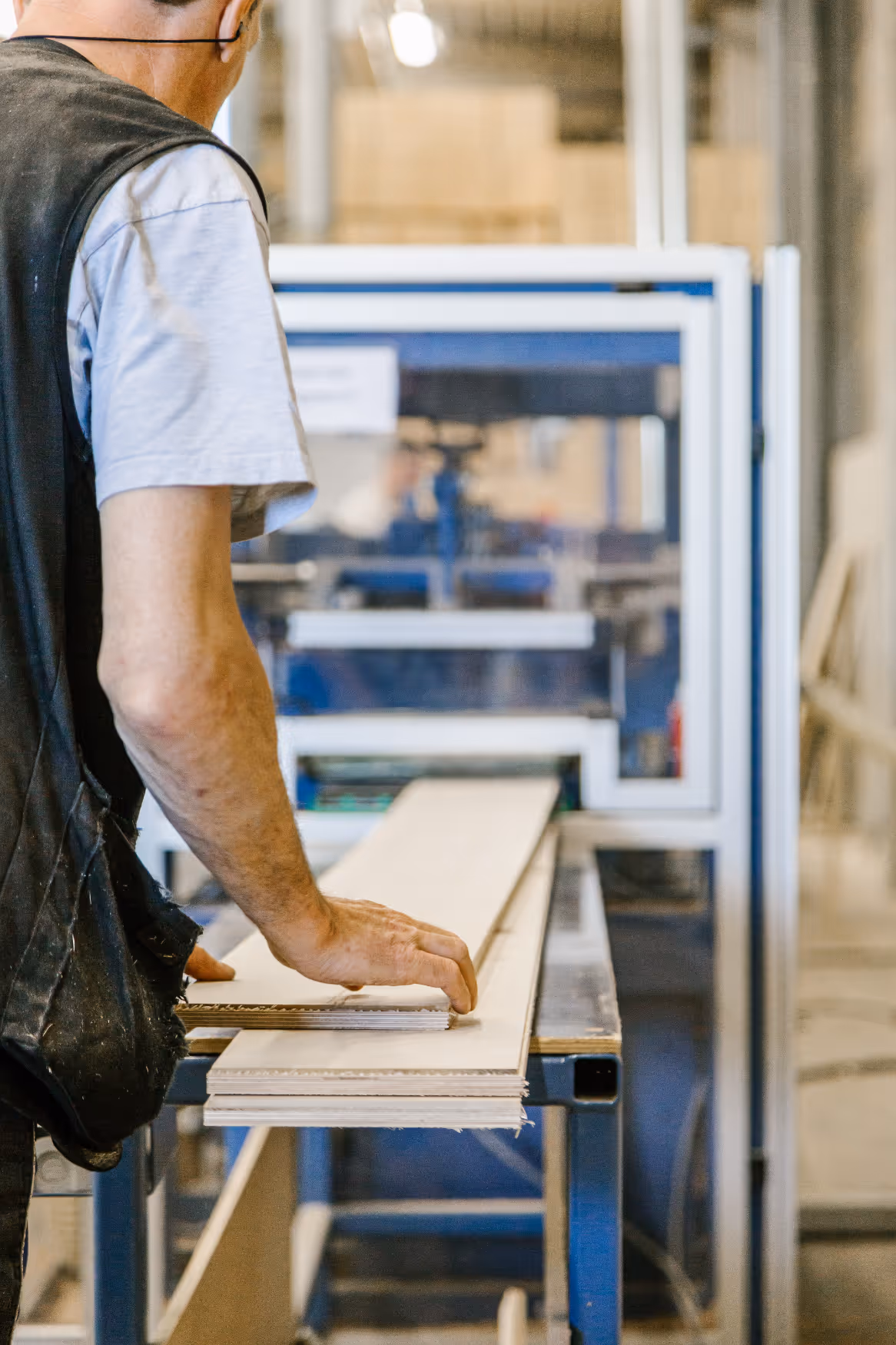 An employee wearing a vest operates a woodworking machine and guides a wooden plank through it. The workshop has an industrial and focused atmosphere.