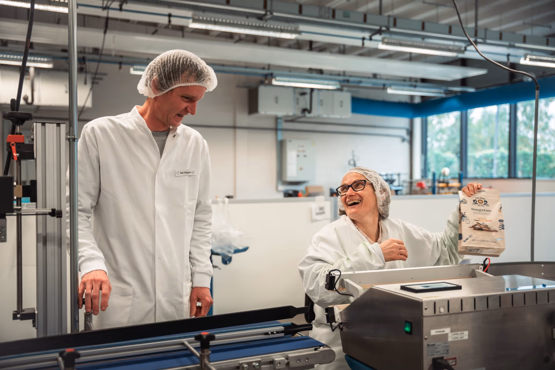 Two people in white lab coats and hairnets smile in a bright factory setting. One of them is holding a packaged product, which conveys a sense of teamwork and achievement.