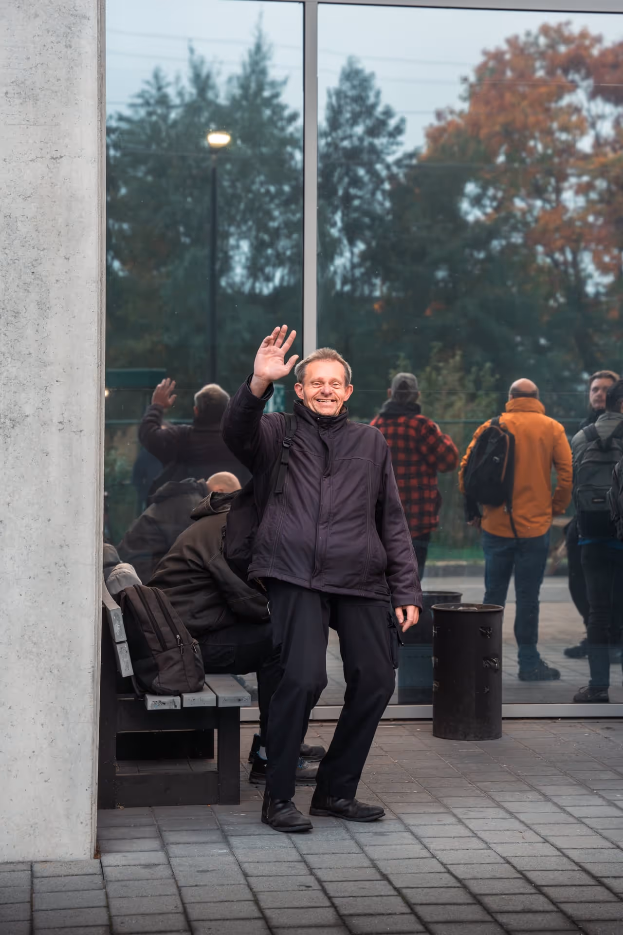 A man in a dark coat waves and smiles in front of a reflective glass wall. Several people with backpacks walk by, with autumn trees in the background.