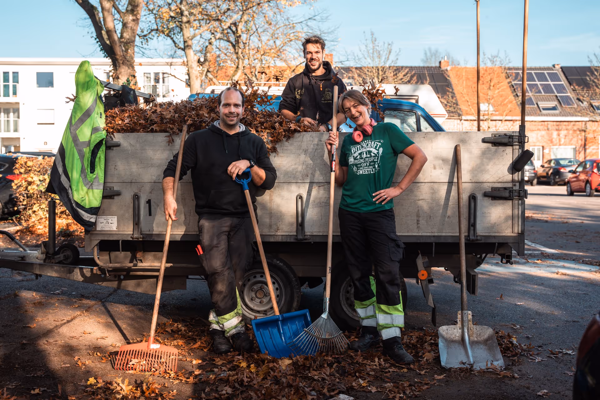Three people are standing smiling in front of a truck full of leaves. Two are holding brooms, the third is sitting on the truck. It is a sunny autumn day, with leaves scattered everywhere.