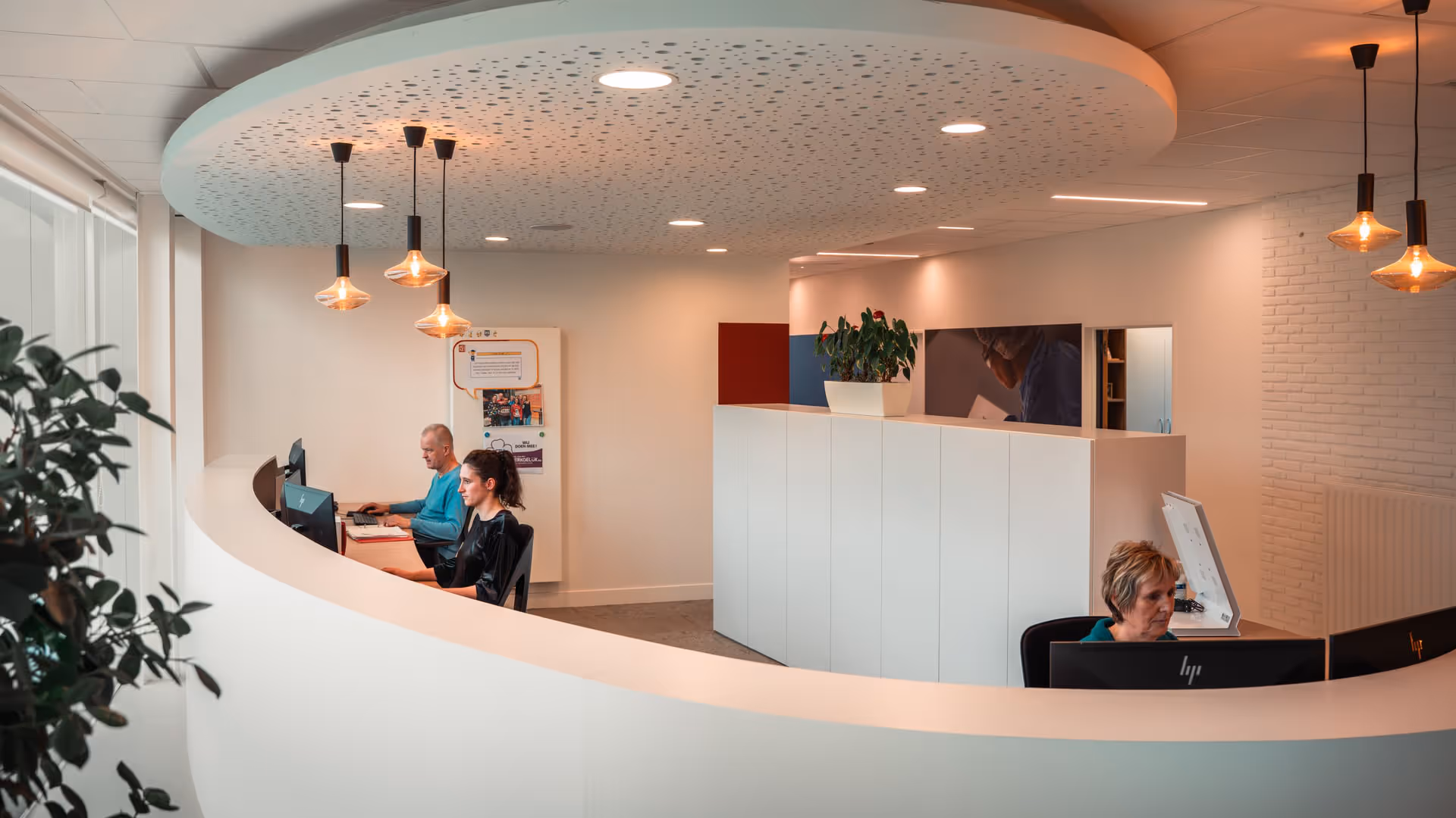 Modern office with a curved white desk and sleek lighting. Three people are working at computers, creating a focused and professional atmosphere.