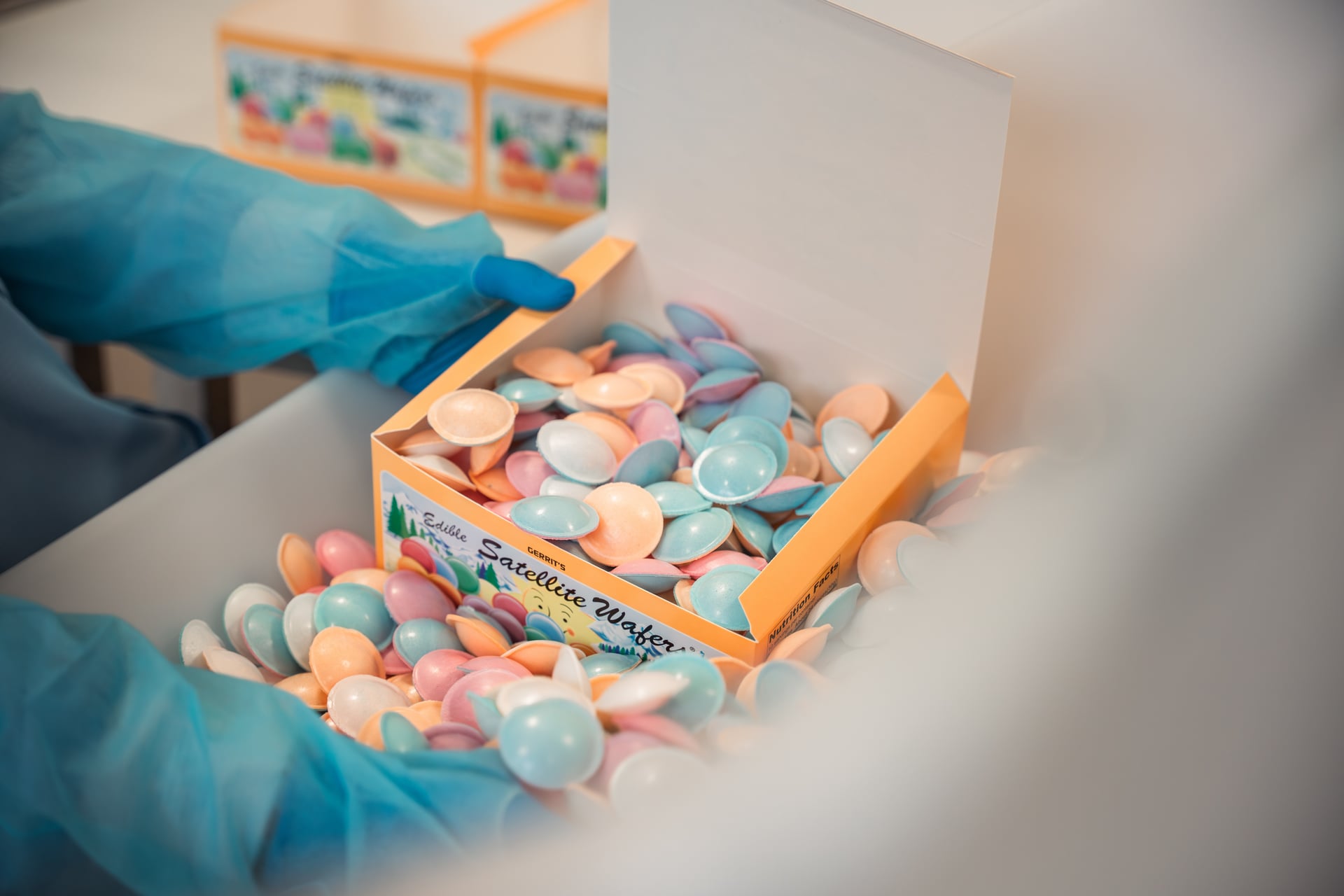 A person wearing blue gloves packs colorful pastel-colored satellite waffles into an orange box. The scene exudes a cheerful, organized production environment.