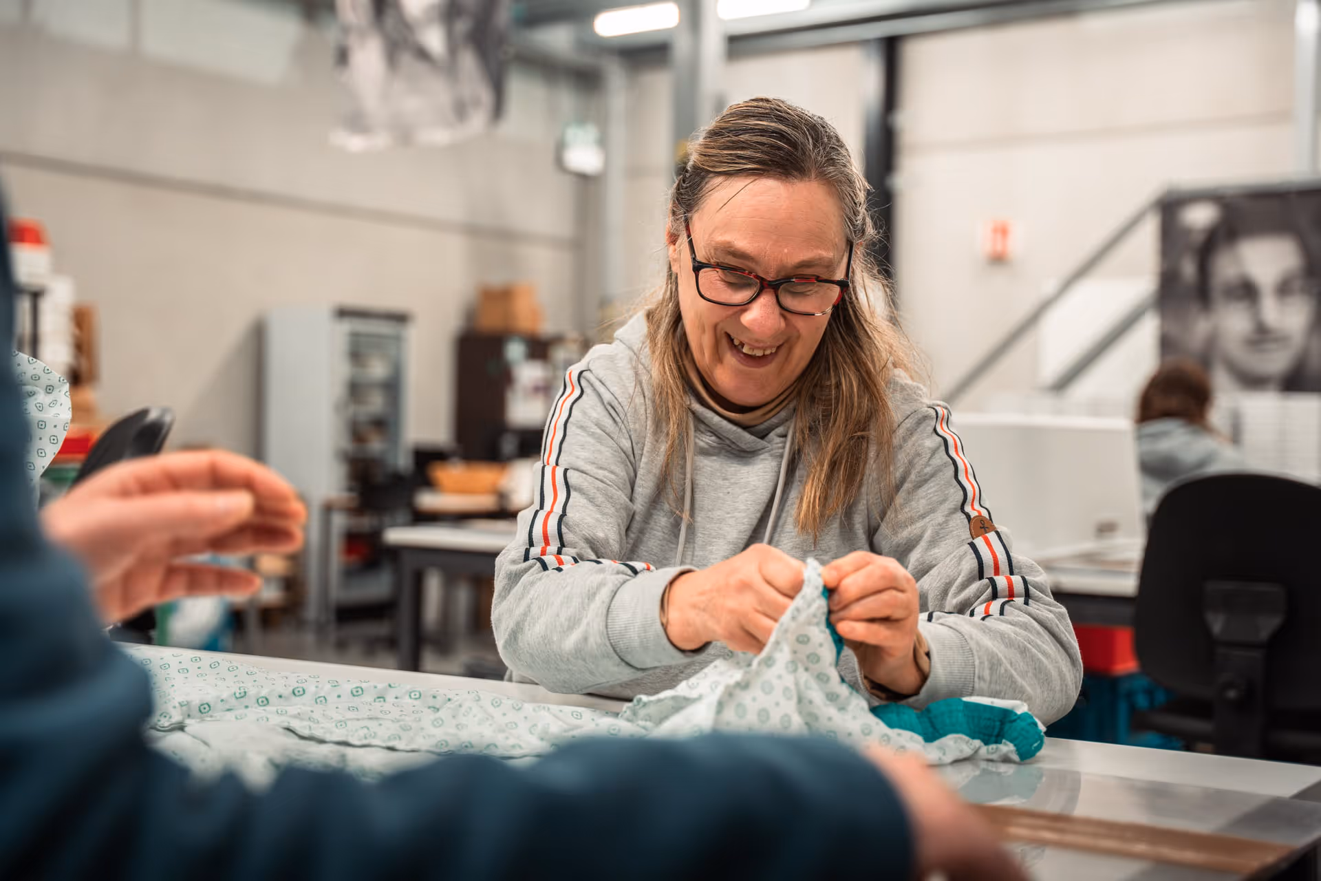 A woman wearing glasses and a gray hoodie is happily sewing fabric in a workshop, concentrating intently on her work. In the background, a workspace with shelves can be seen.