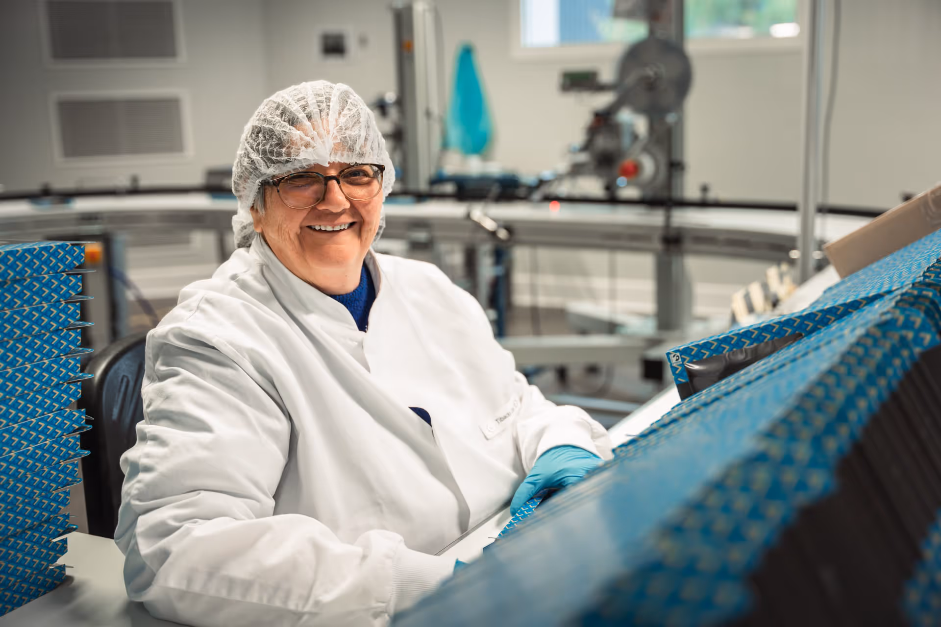 A smiling factory worker in a white coat and hairnet sits next to a conveyor belt full of blue boxes, radiating professionalism and satisfaction.