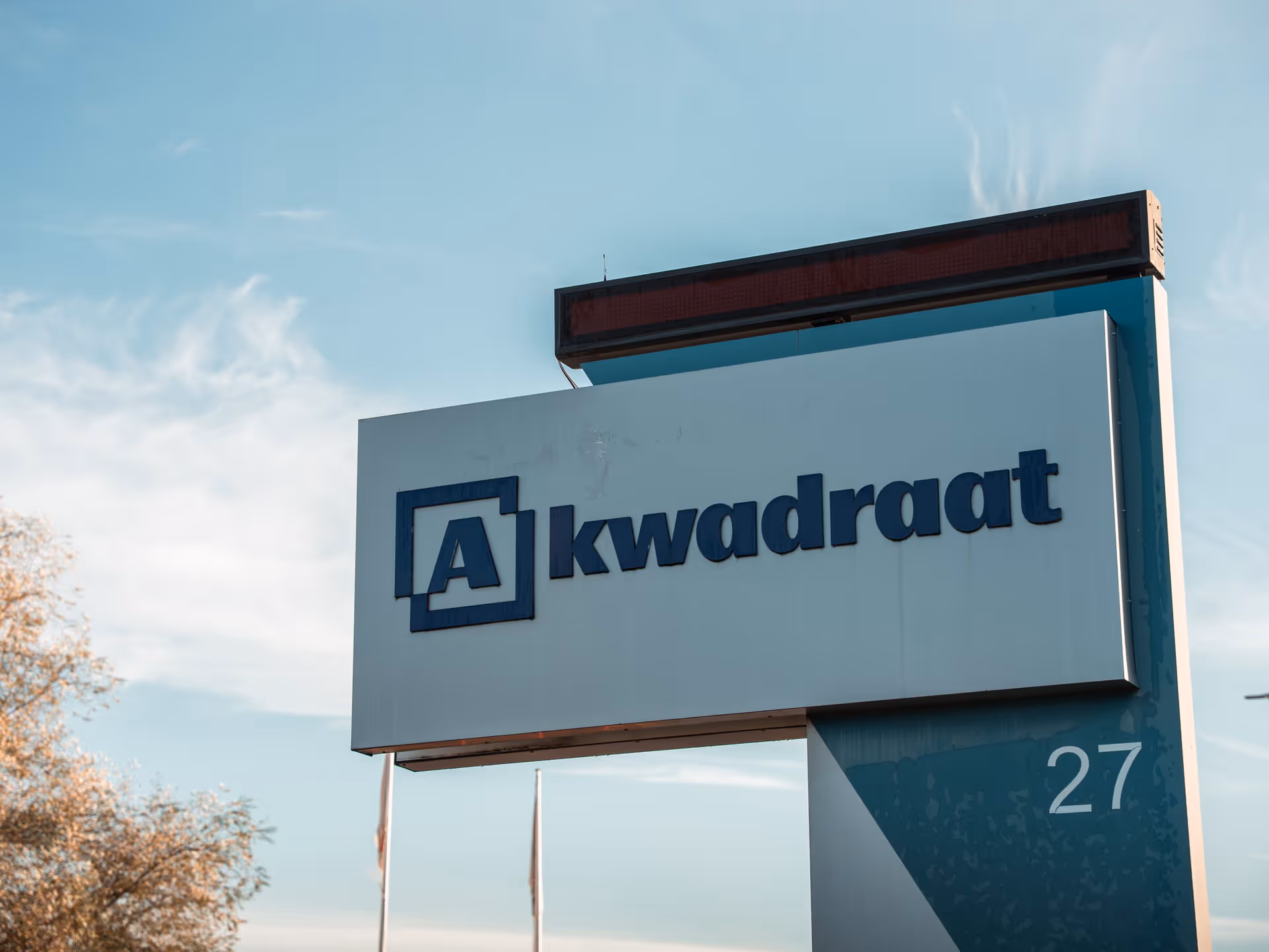 Sign with "A-kwadraat" and logo against a bright blue sky. The number 27 is visible on the stand, giving it a professional, businesslike appearance.
