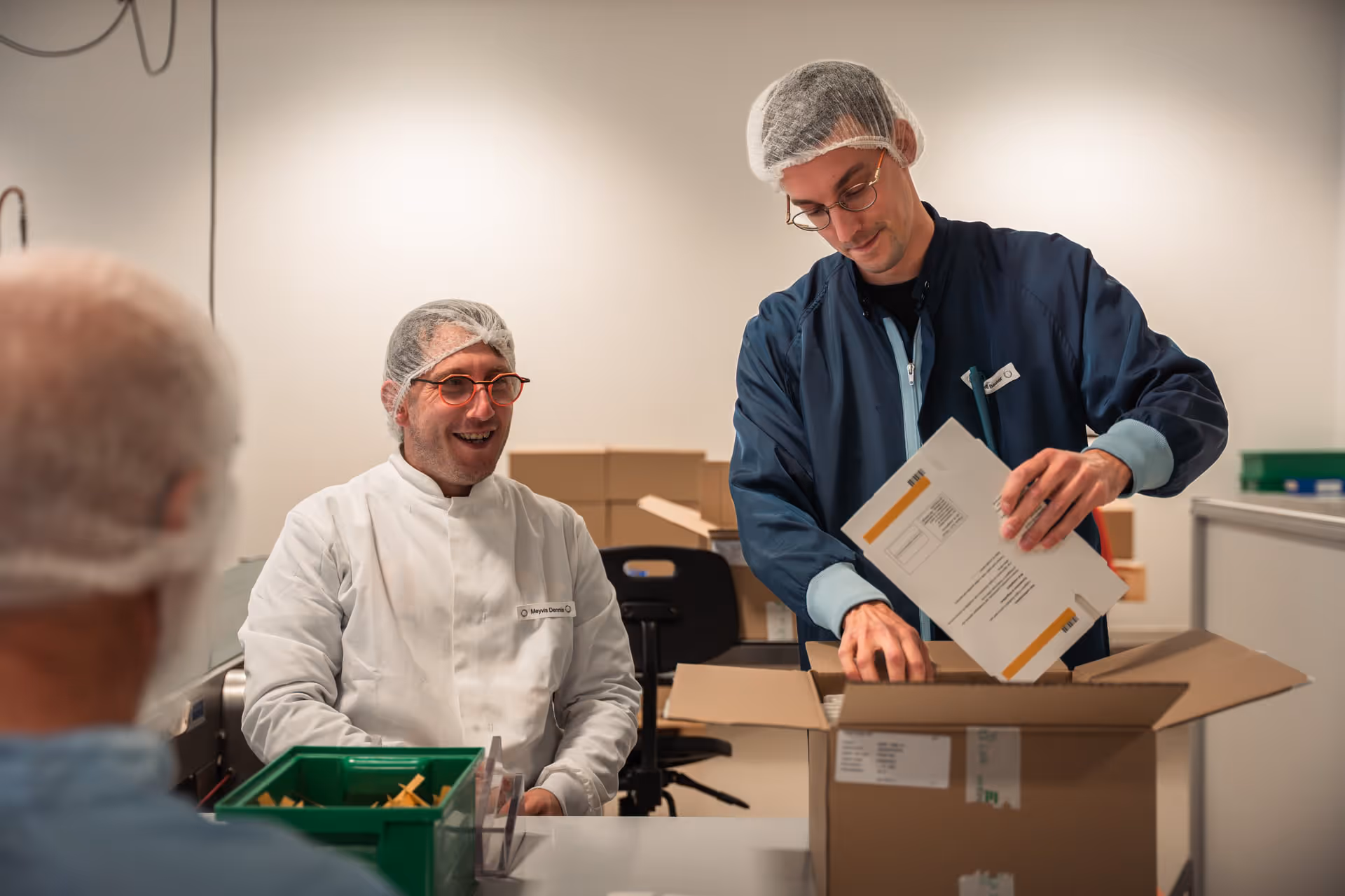Two people wearing hairnets and lab coats are packing boxes in a well-lit room. One is sitting down, smiling, while the other is standing and working intently.