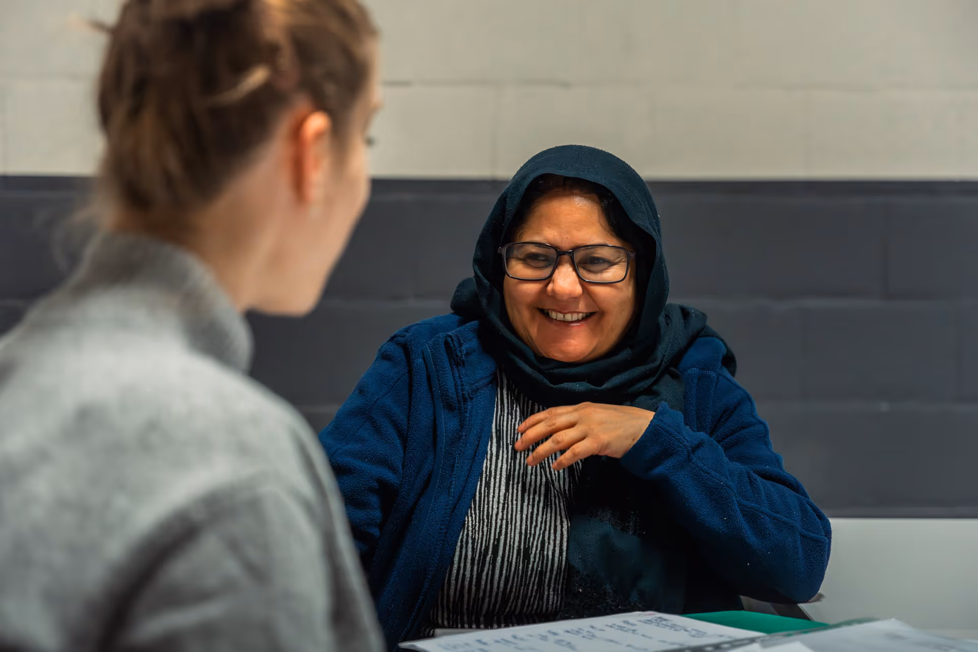 A smiling woman wearing a blue headscarf and glasses is talking to someone across the table. The setting is warm and inviting, suggesting a positive interaction.