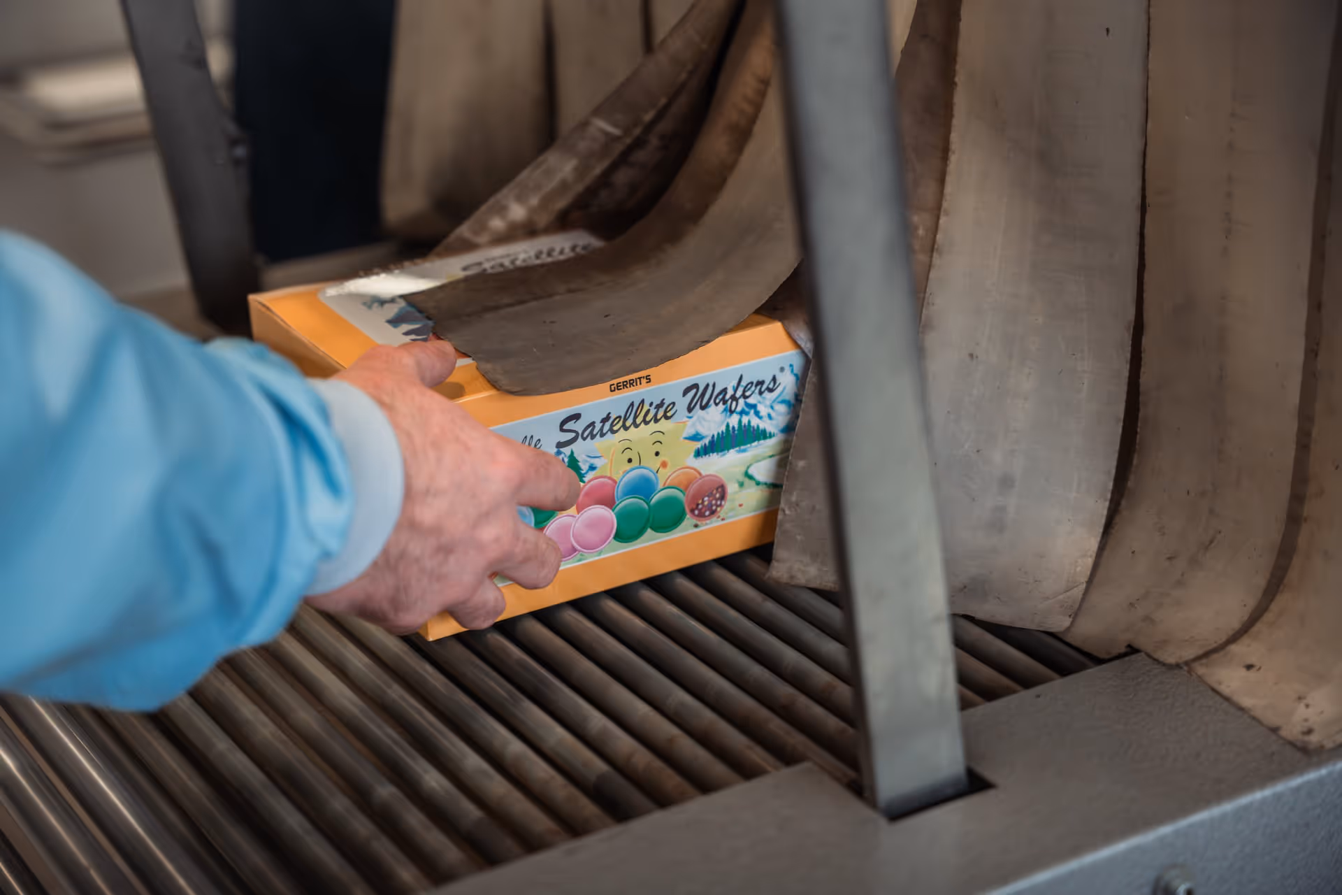 A person in a blue sleeve places a box of "Satellite Wafers" on a metal conveyor belt. The box is colorful, with images of planets and a smiling sun.