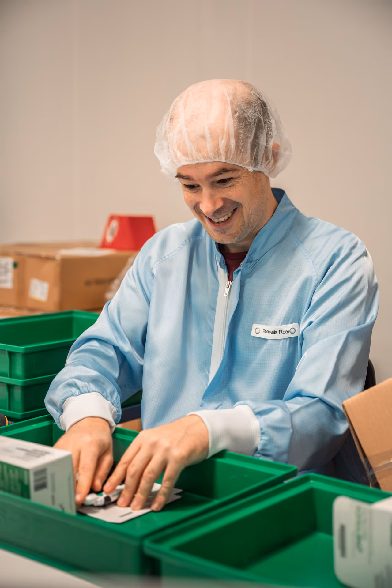 A smiling man in a blue lab coat and hairnet at a workstation, sorting boxes and items into green bins, radiating concentration and satisfaction.