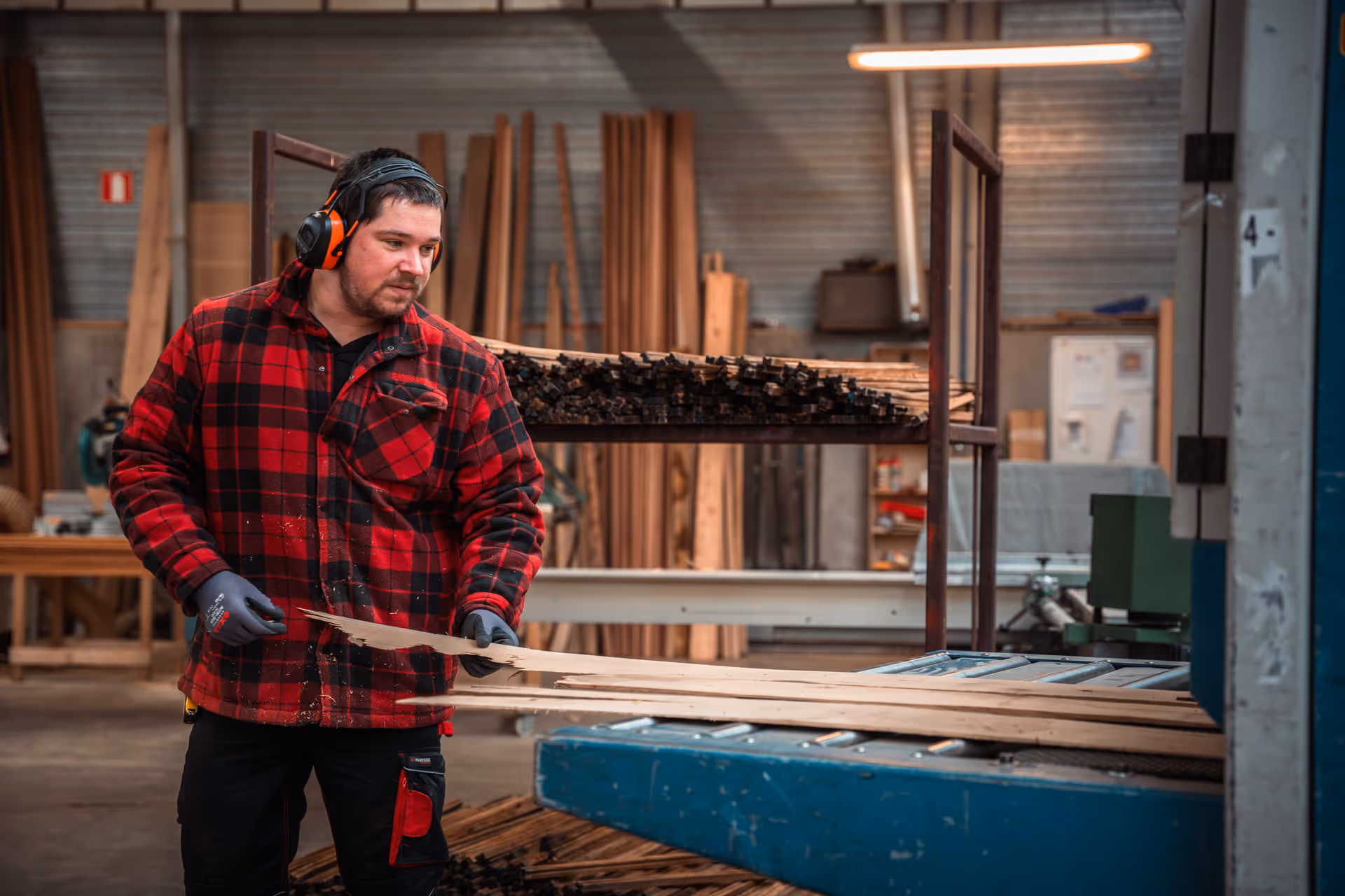 A man wearing a red checked jacket and earmuffs is working in a woodworking shop, processing wooden planks with a machine. The workshop is well-organized yet busy.