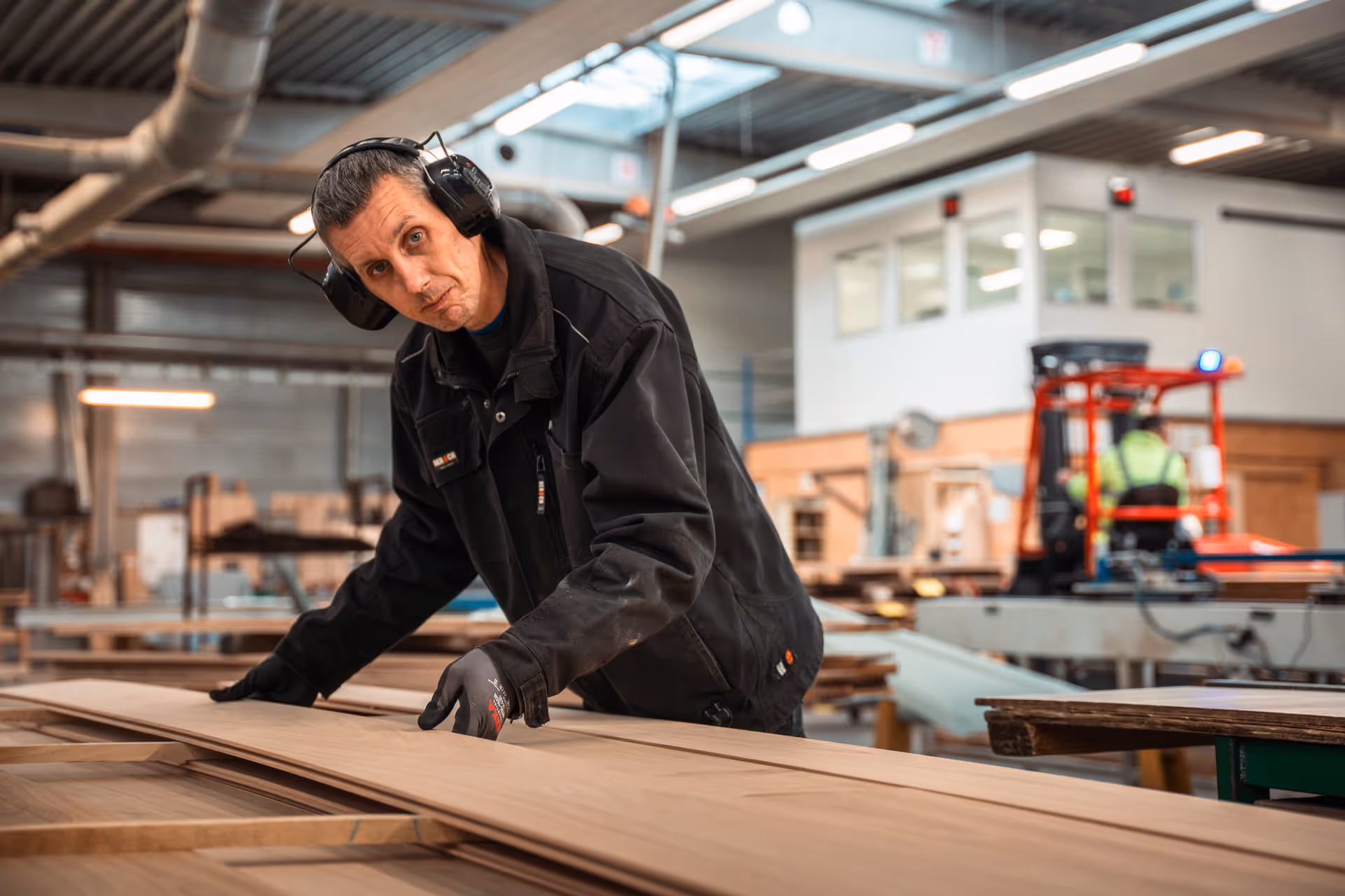 A man wearing ear protection and gloves inspects a wooden panel in a workshop. The setting is industrial, with a focused and industrious atmosphere.