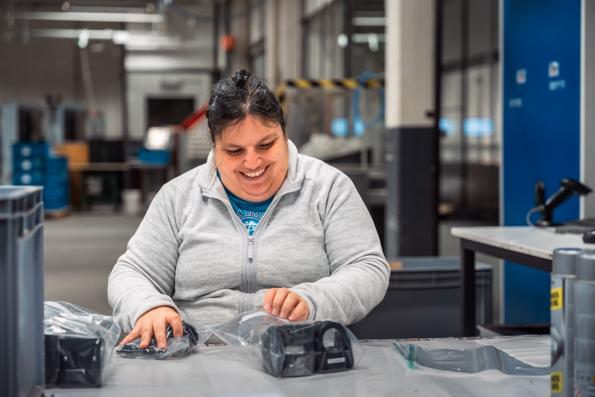 A woman in a gray sweater smiles as she packs items into plastic bags in a bright, well-organized workspace, exuding a sense of satisfaction and productivity.