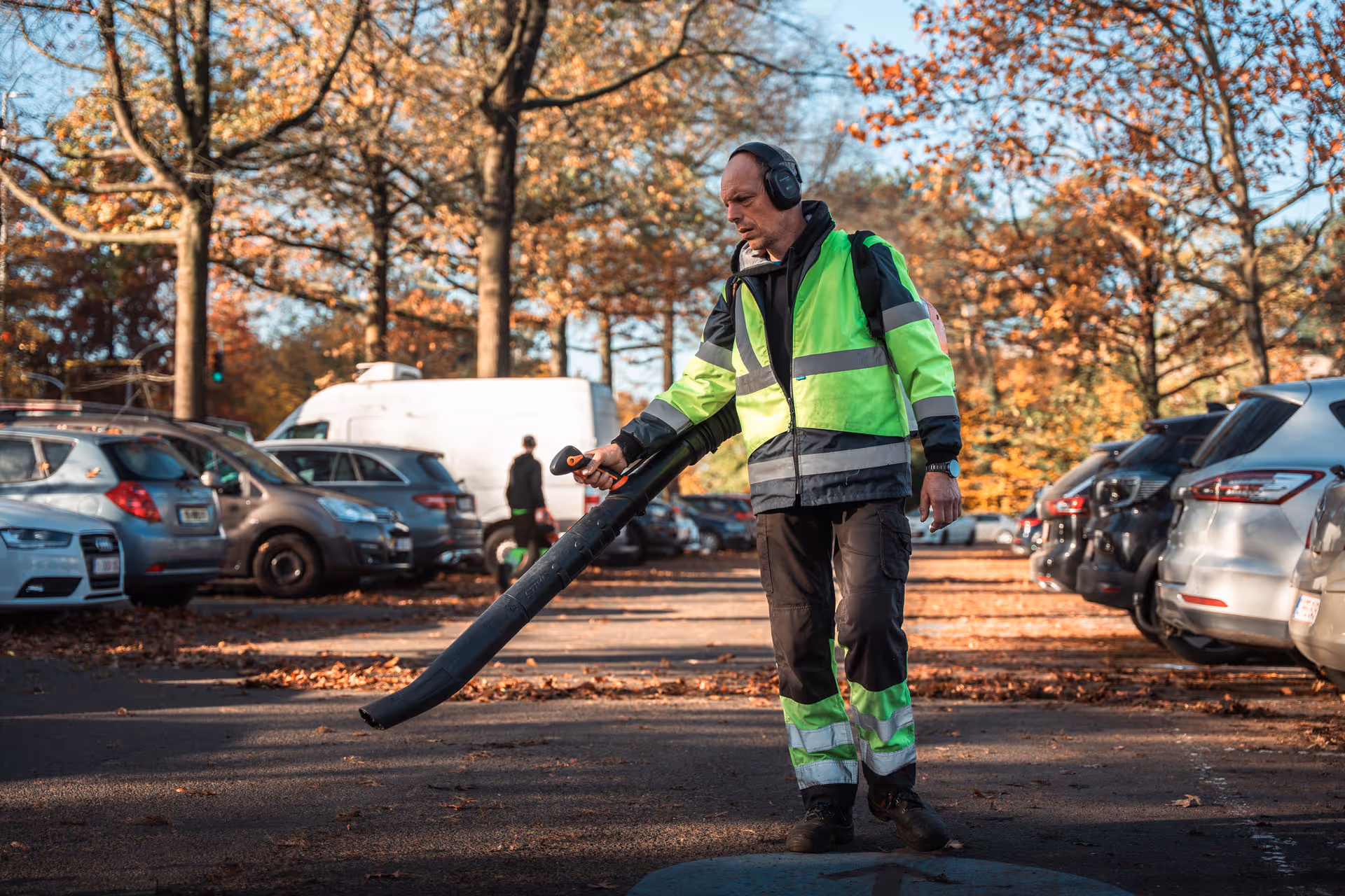 An employee wearing a fluorescent jacket uses a leaf blower in a parking lot. Autumn leaves cover the ground. Parked cars and bare trees are visible in the background.