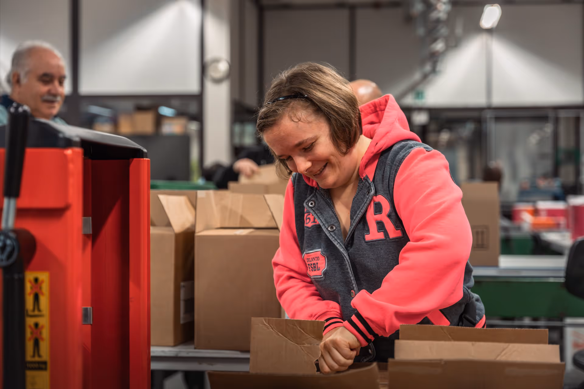A woman in a pink and gray hoodie smiles as she packs cardboard boxes in a warehouse. The atmosphere is busy and positive.
