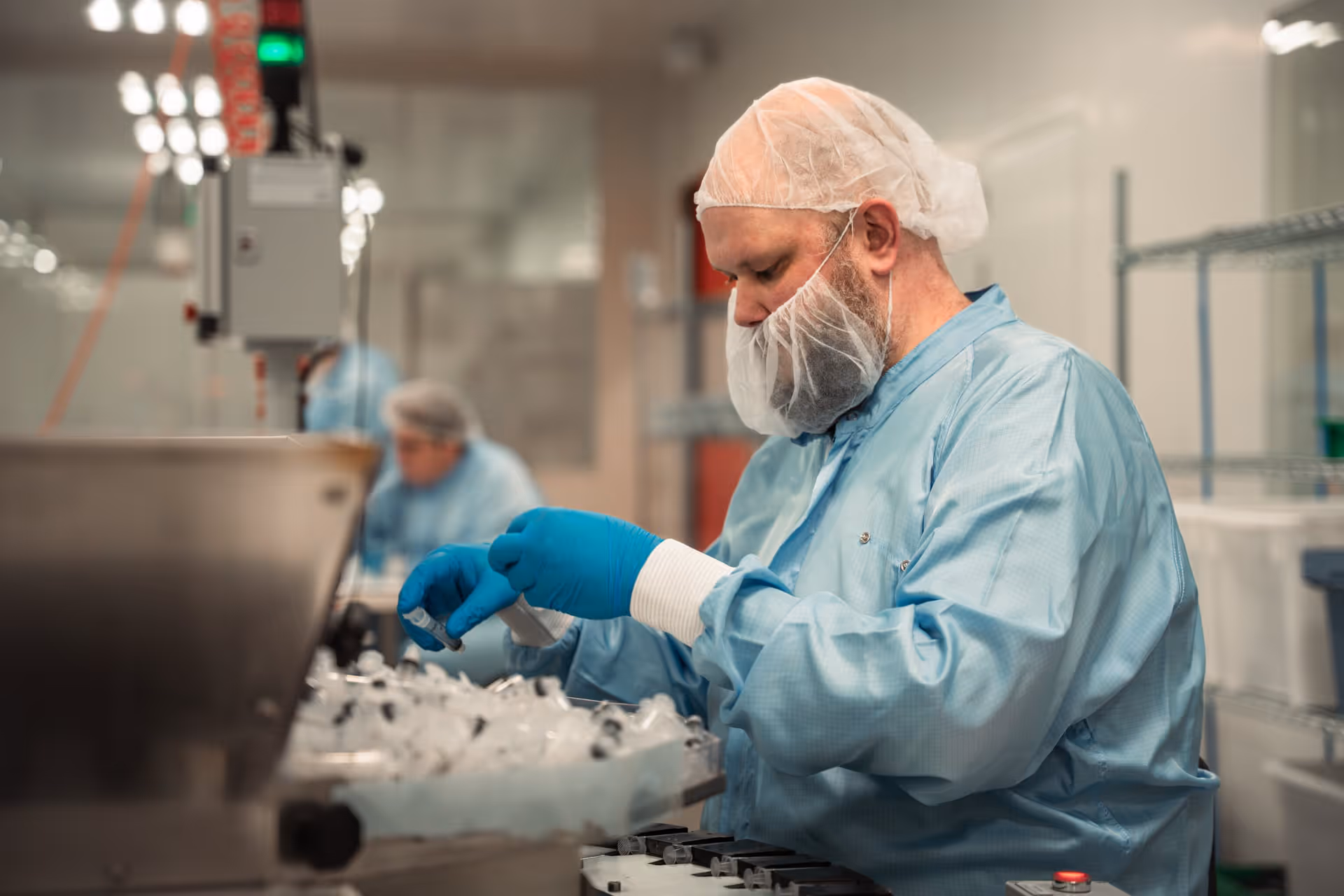 A person wearing protective clothing, including a hairnet and gloves, works intently in a cleanroom environment and handles laboratory equipment. The environment is clinical and focused.