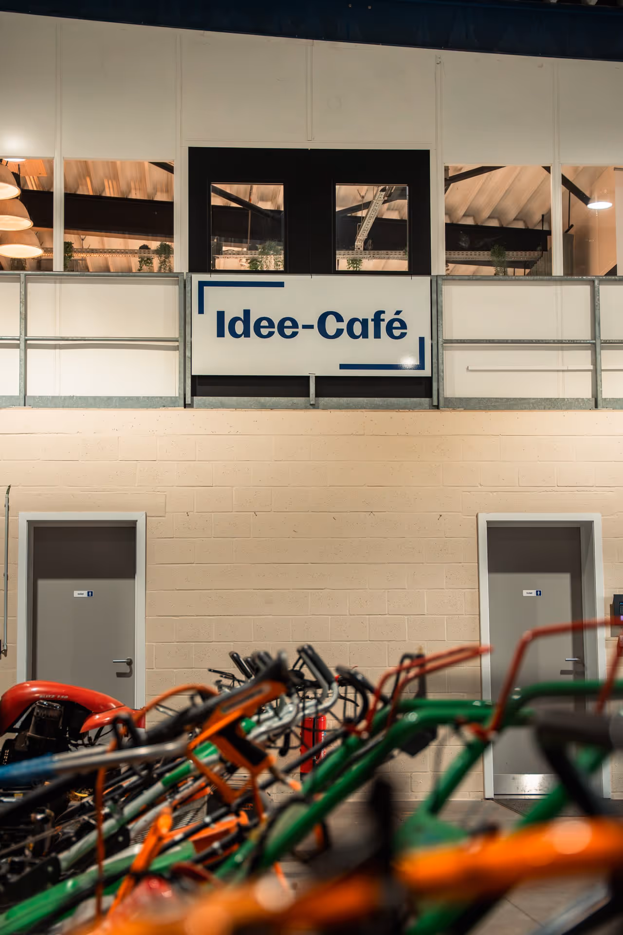 Interior of a warehouse-style space with a sign reading "Idee-Café" above two closed doors. In the foreground, bicycles with colorful frames are parked.