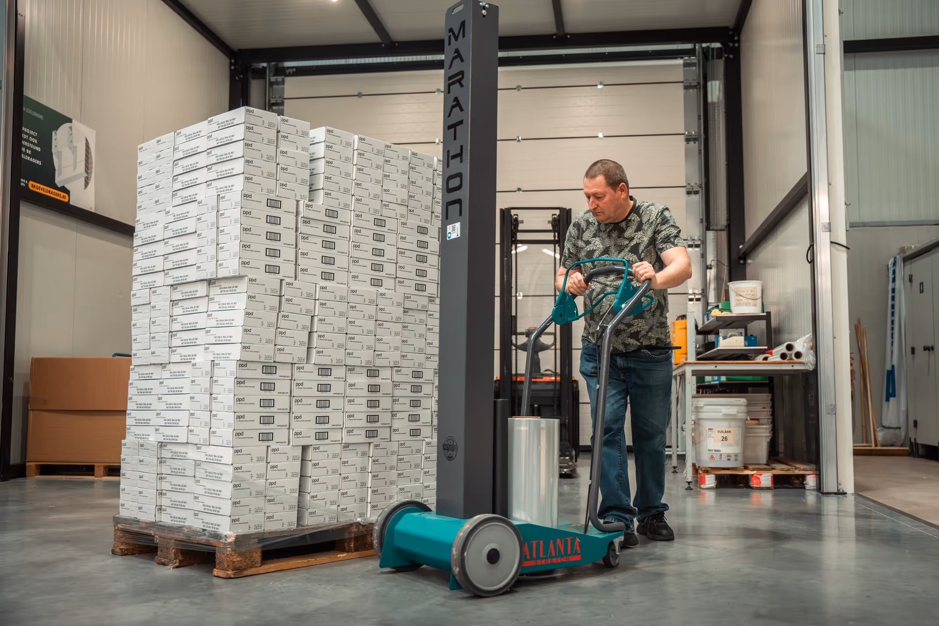 A man operates a blue-green pallet wrapper next to a tall stack of white boxes on a wooden pallet in a spacious, well-lit warehouse.