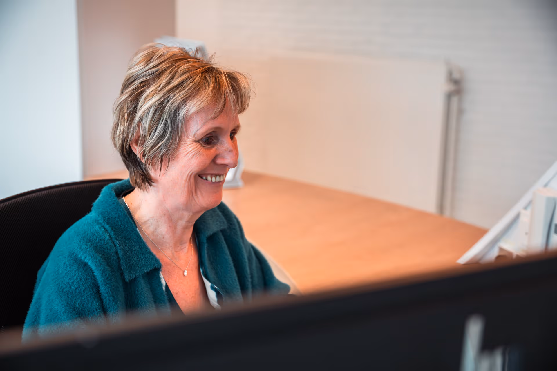 A woman with short hair and a green sweater smiles as she looks at her computer in a bright office. The setting exudes a positive, focused atmosphere.