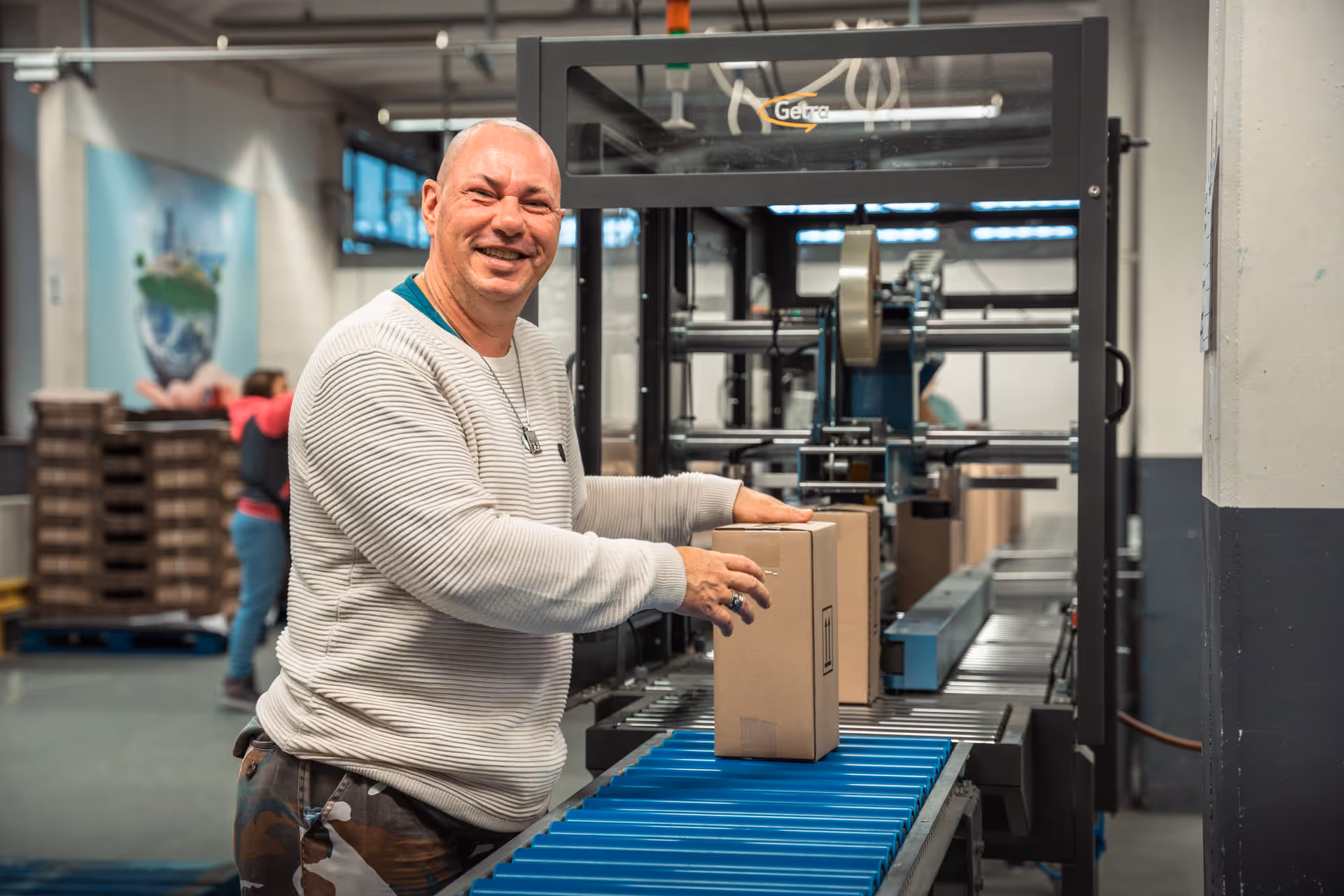 A smiling man in a factory setting places a cardboard box on a conveyor belt. The atmosphere is industrial, with a warm and positive tone.