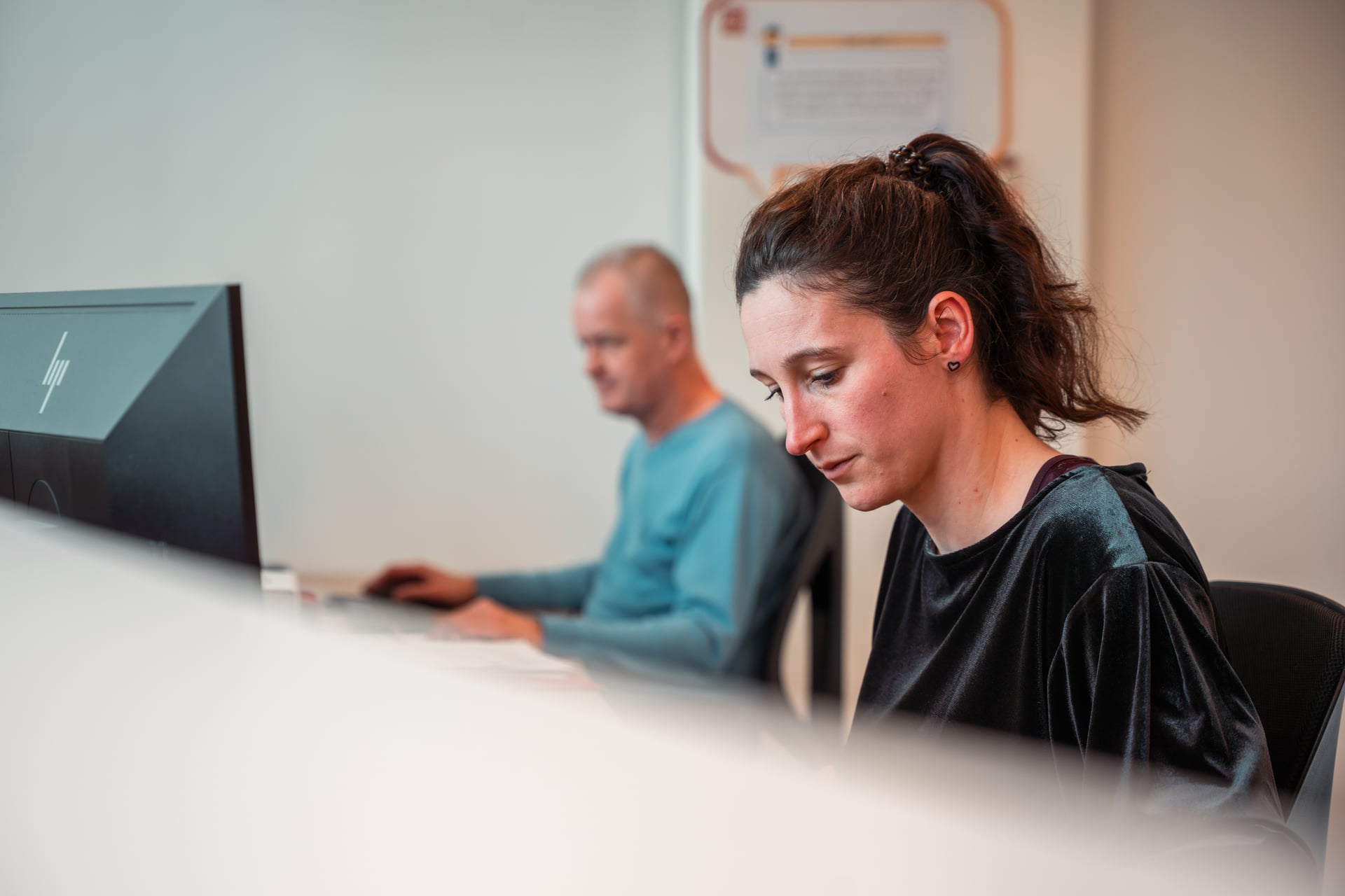 Three vertical flags on poles against a bright blue sky. The flags have diagonal stripes in green, yellow, and red, with stylized text and a logo.