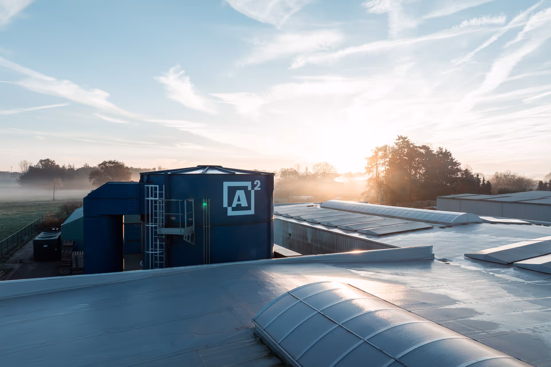 View from the roof of an industrial building at sunrise. Sunlight gently shines through the trees in the background, with a blue silo bearing the inscription "A²" clearly visible.