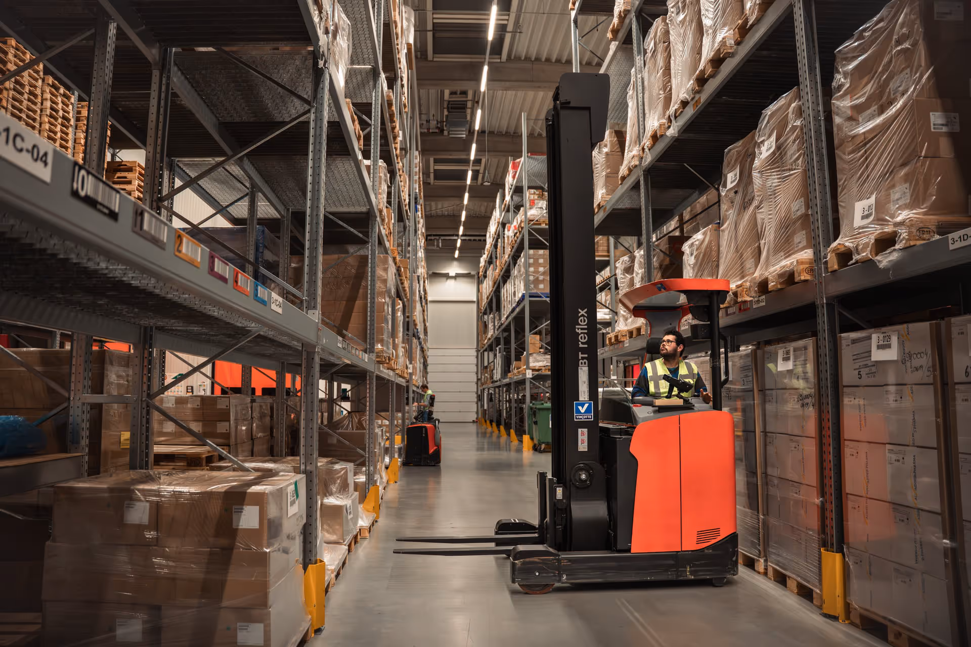 A warehouse worker driving a red and black forklift truck in an aisle filled with stacked pallets and boxes, under bright overhead lighting.