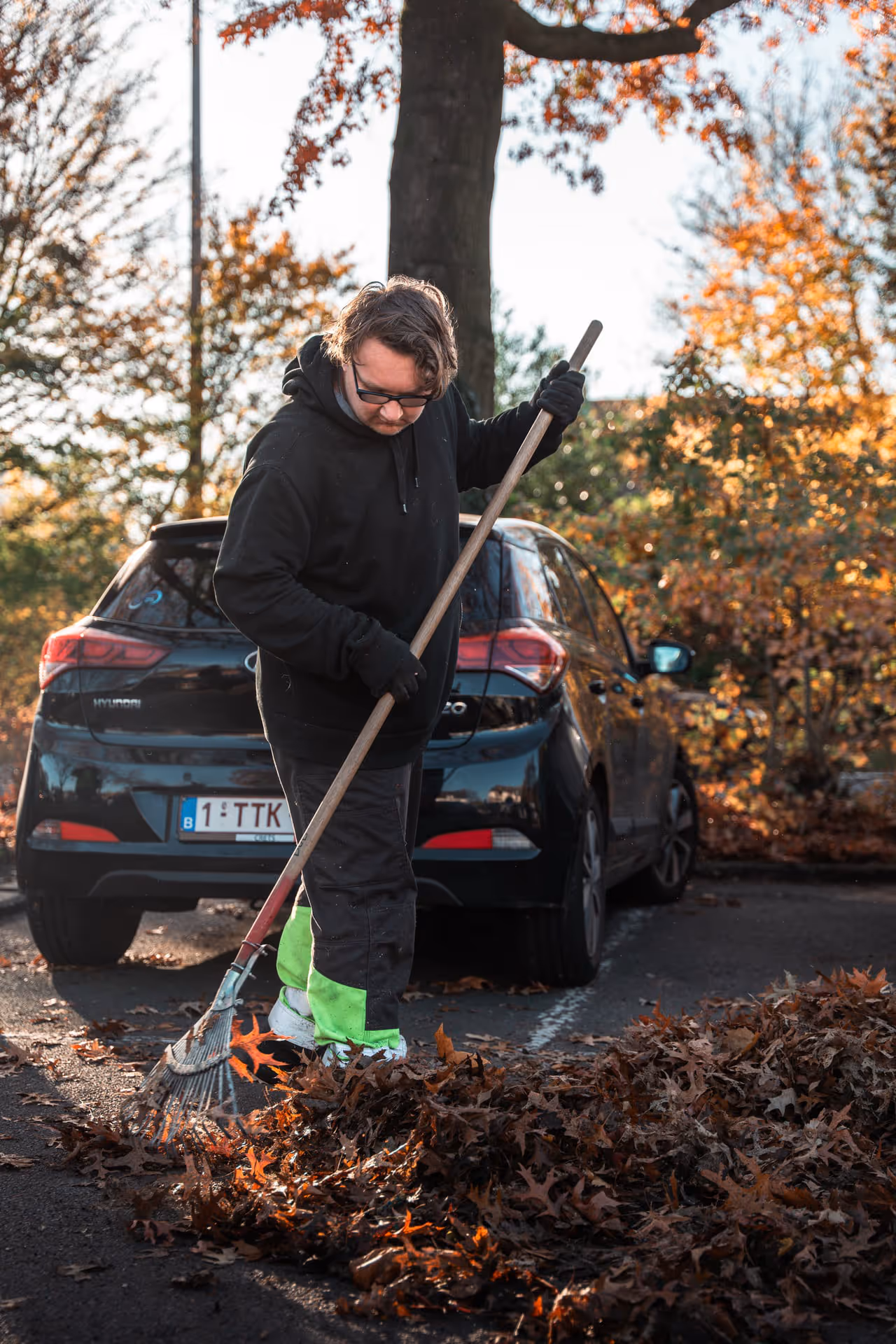 Person raking autumn leaves next to a parked car, wearing a black hoodie and gloves. The scene takes place in a sunny autumn setting with colorful foliage.