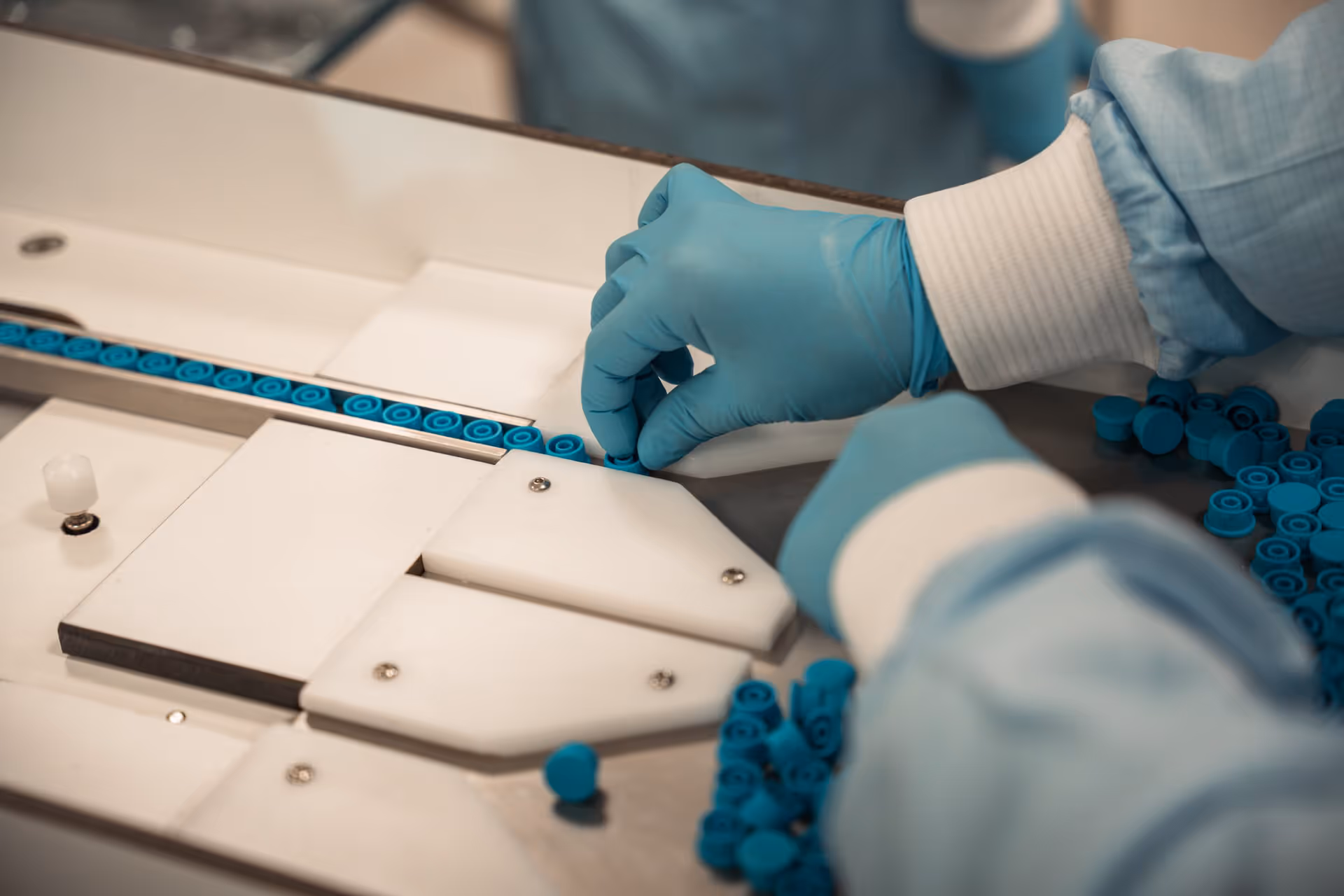 Gloves in blue surgical clothing organize blue caps on a clean conveyor belt, which exudes precision and cleanliness in a sterile production environment.
