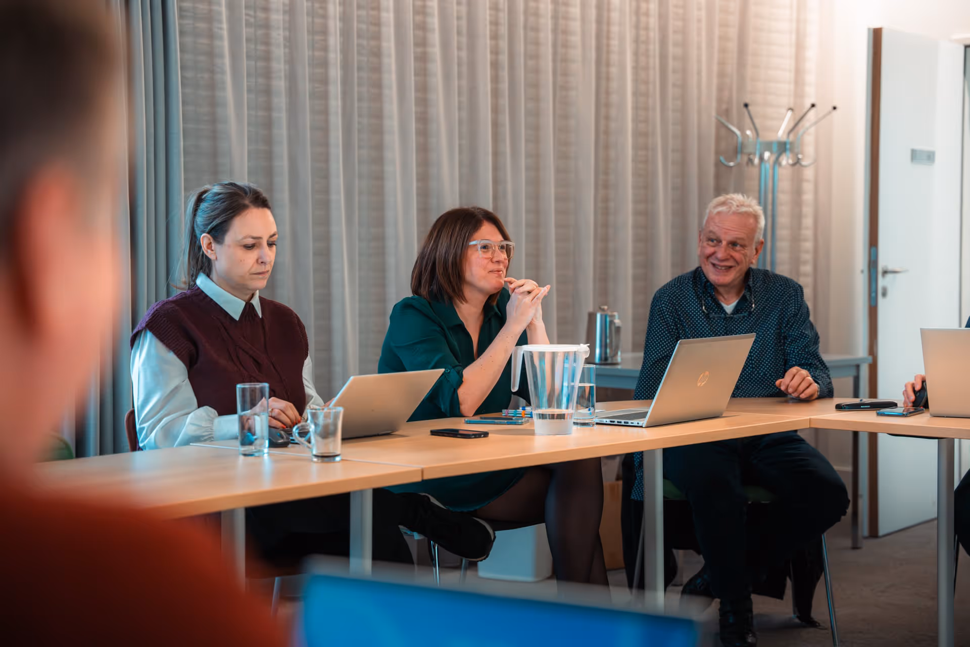 Three people are sitting at a conference table with laptops and are engaged in conversation. The atmosphere is focused and professional, with neutral-colored curtains in the background.