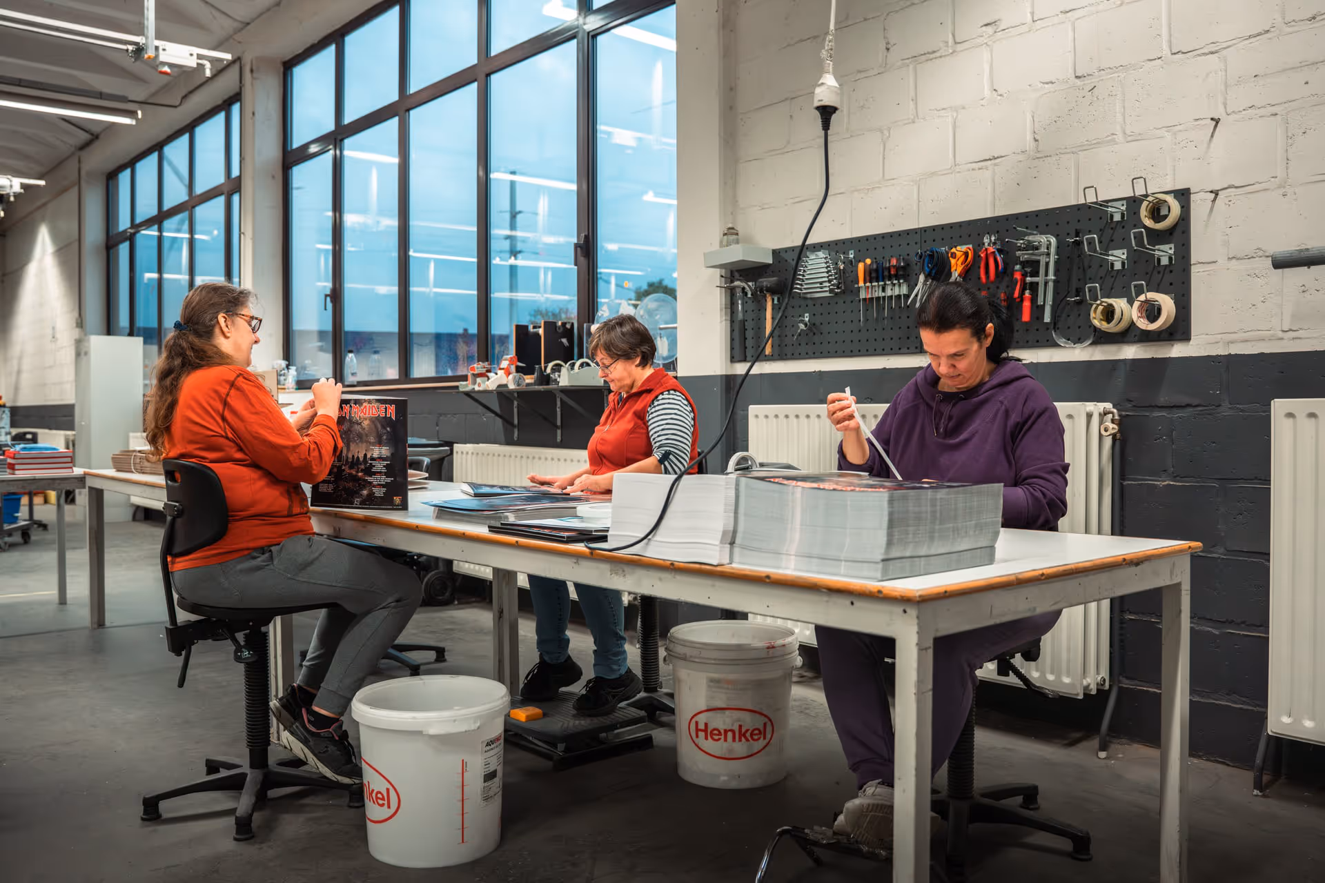 Three people are busy putting together printed material at a table in a bright workspace. Behind them, tools hang on a pegboard, conveying a sense of focus and collaboration.