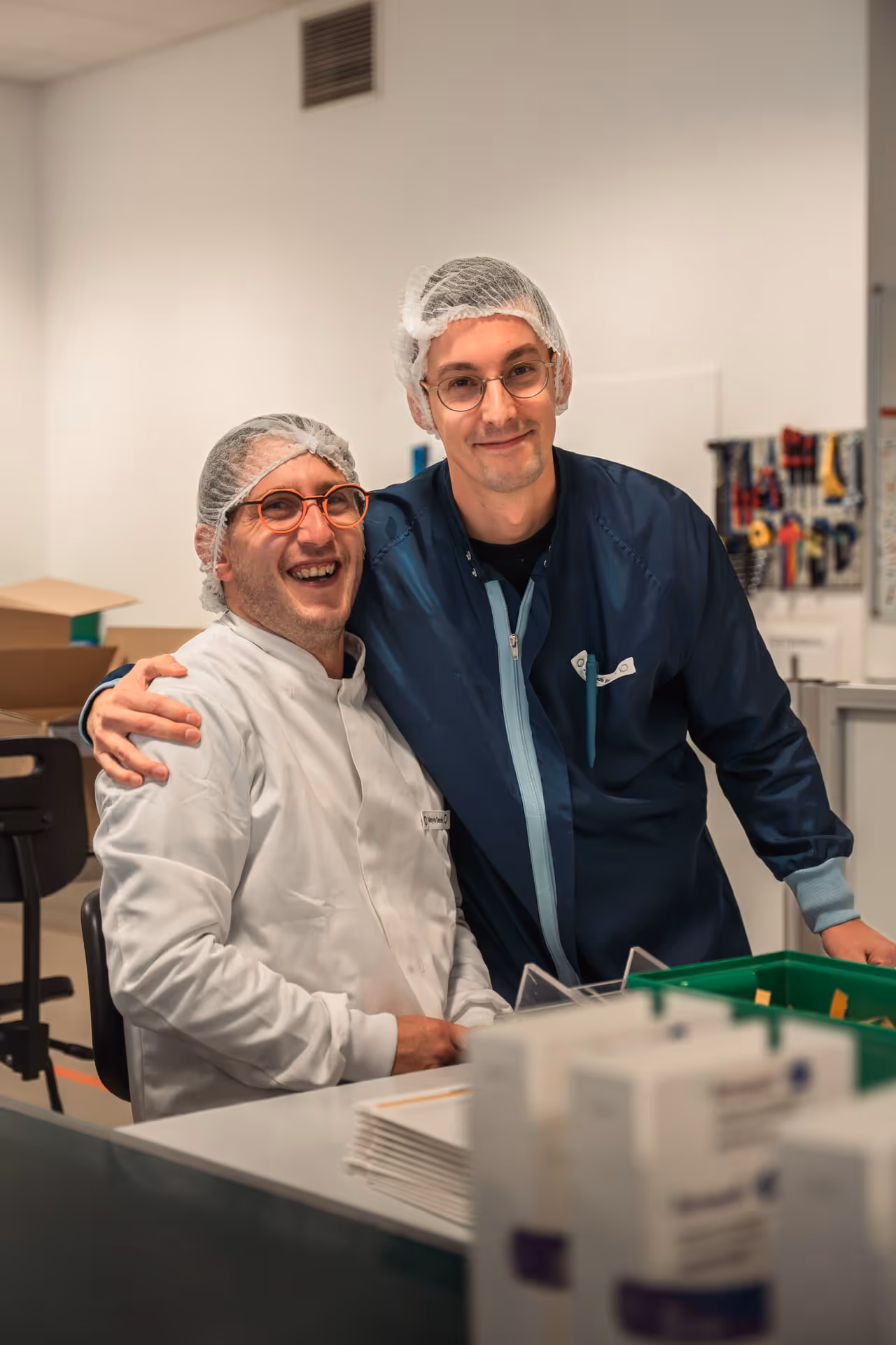 Two men in a laboratory, wearing hairnets and glasses, smile warmly at the camera. One is seated, the other is standing with an arm around him. The atmosphere is pleasant.