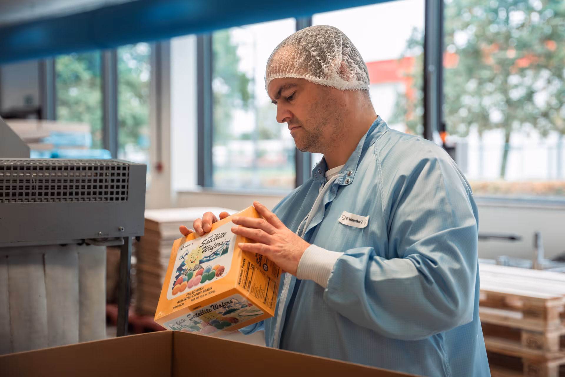 A man in a blue lab coat and hairnet inspects a colorful candy box in a factory. Large windows offer a view of green surroundings. Concentrated atmosphere.