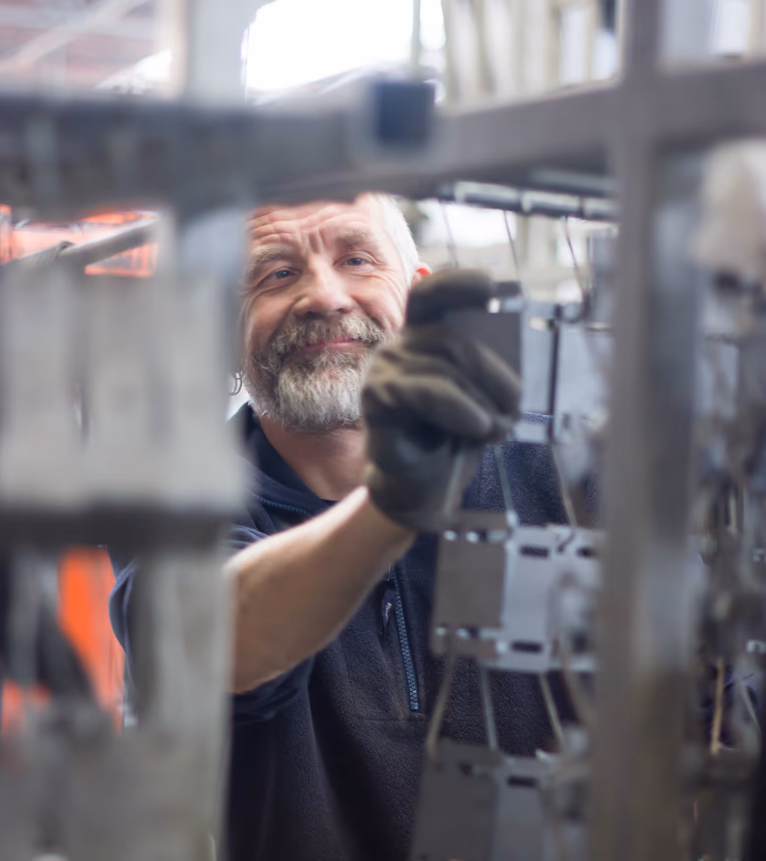 Smiling man with a beard working in a factory, wearing gloves and holding a metal frame. The surroundings have an industrial feel and exude craftsmanship.