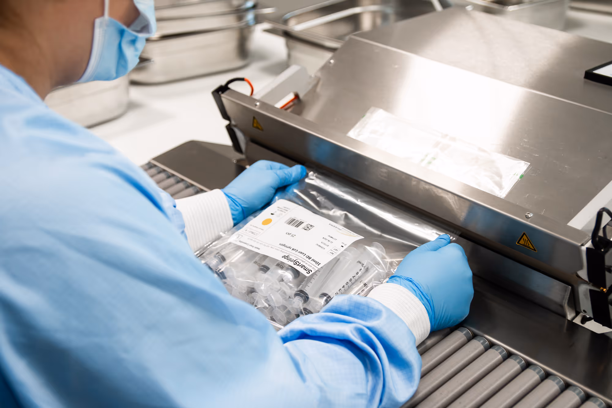 A healthcare provider in blue surgical scrubs and gloves seals medical supplies in a sterilization bag. The environment is clean and professional.