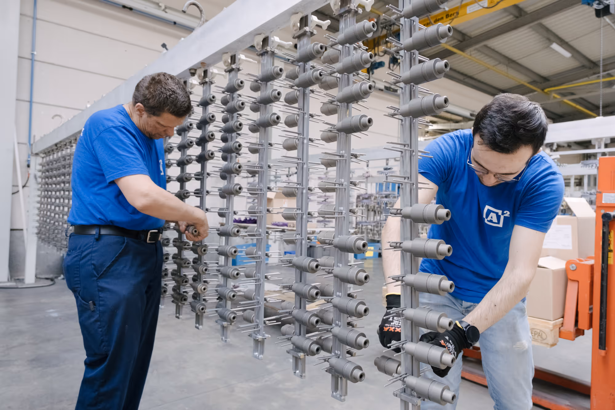 Two workers in blue shirts are adjusting machines in a factory. Metal parts are arranged in rows, creating a focused and industrious atmosphere.