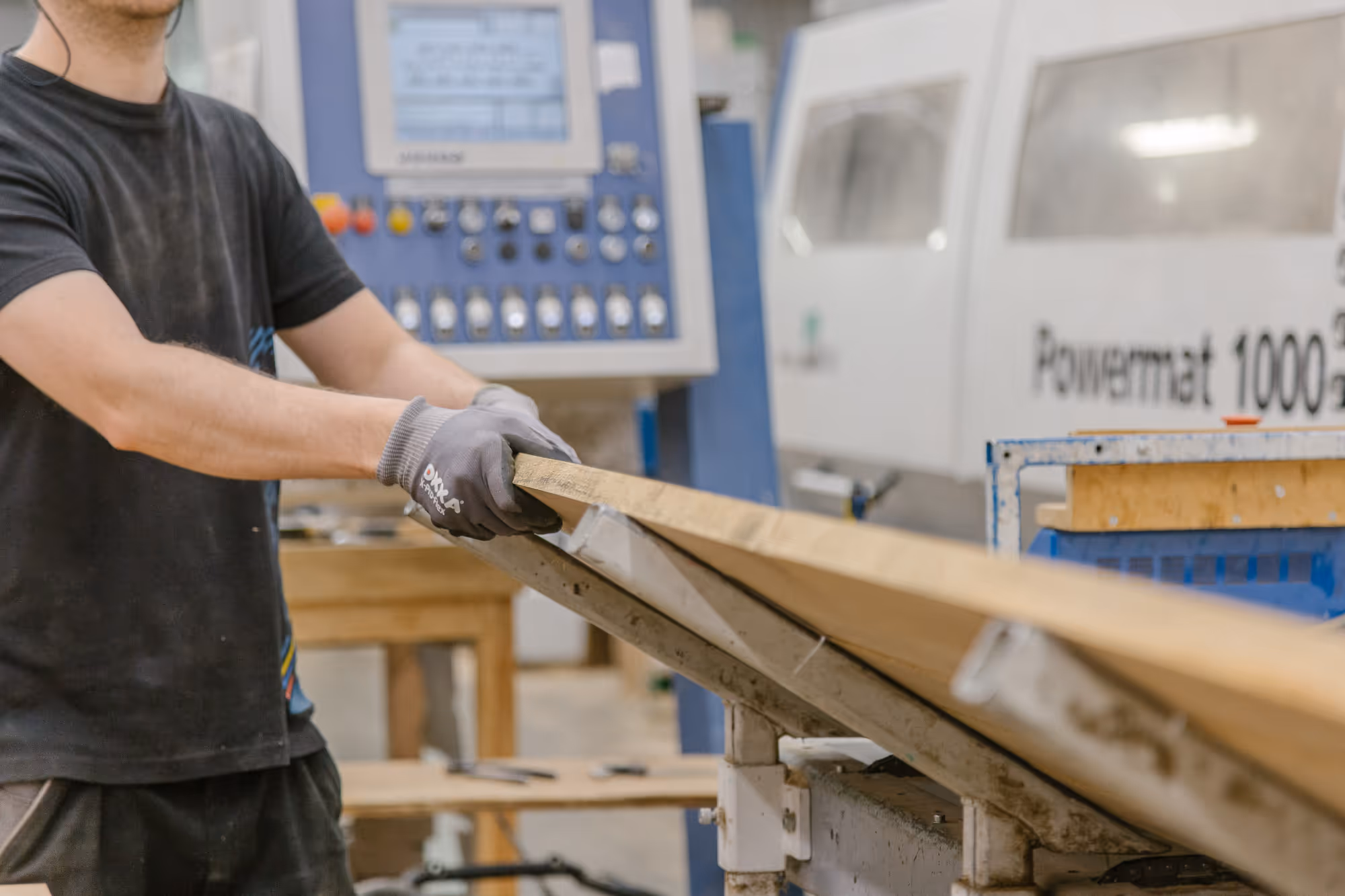 A person wearing gloves operates a woodworking machine and adjusts a wooden plank. Industrial equipment and a monitor can be seen in the background. The setting is a busy workshop.