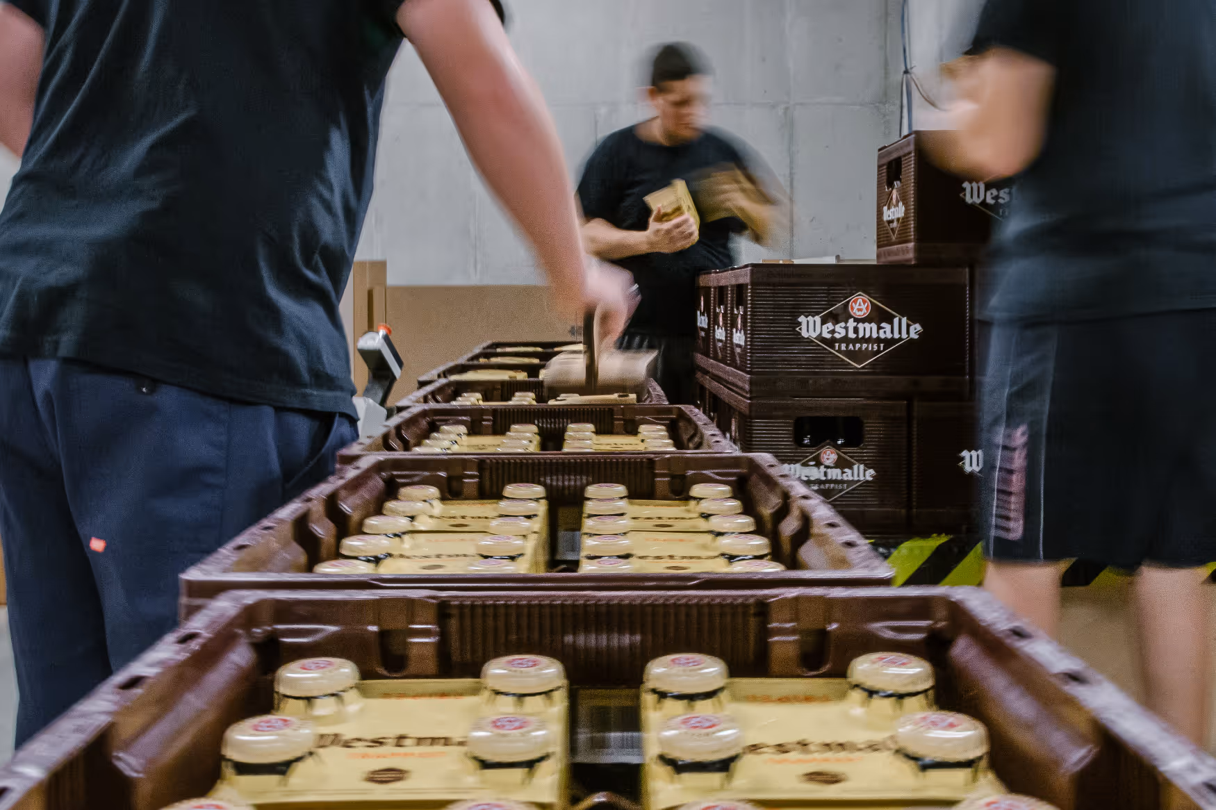 People from A-kwadraat stack crates of Westmalle Trappist beer in a warehouse or brewery environment.