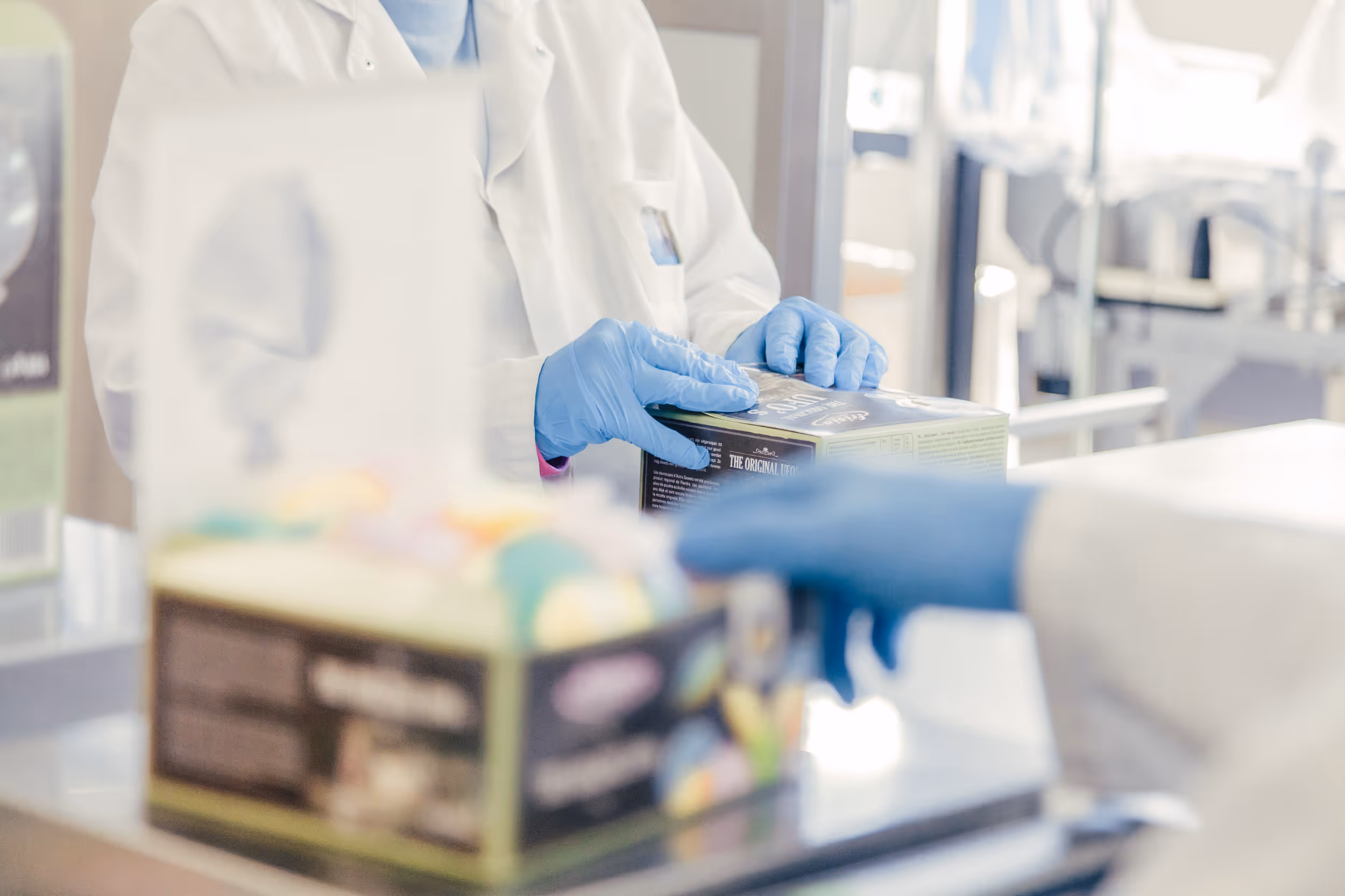 Two people wearing lab coats and blue gloves handle packaged products in a bright, clinical environment, suggesting a sterile working environment.