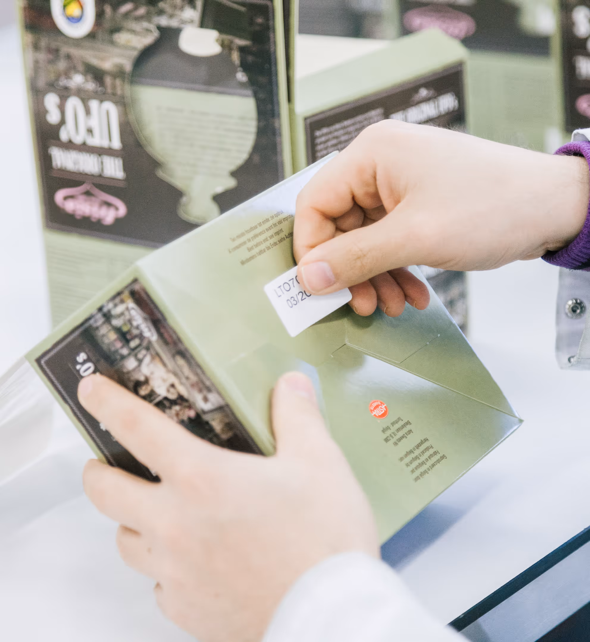 Two hands stick a sticker with partially visible text onto a green product box. Other similar boxes are visible in the background. The focus is on the packaging.