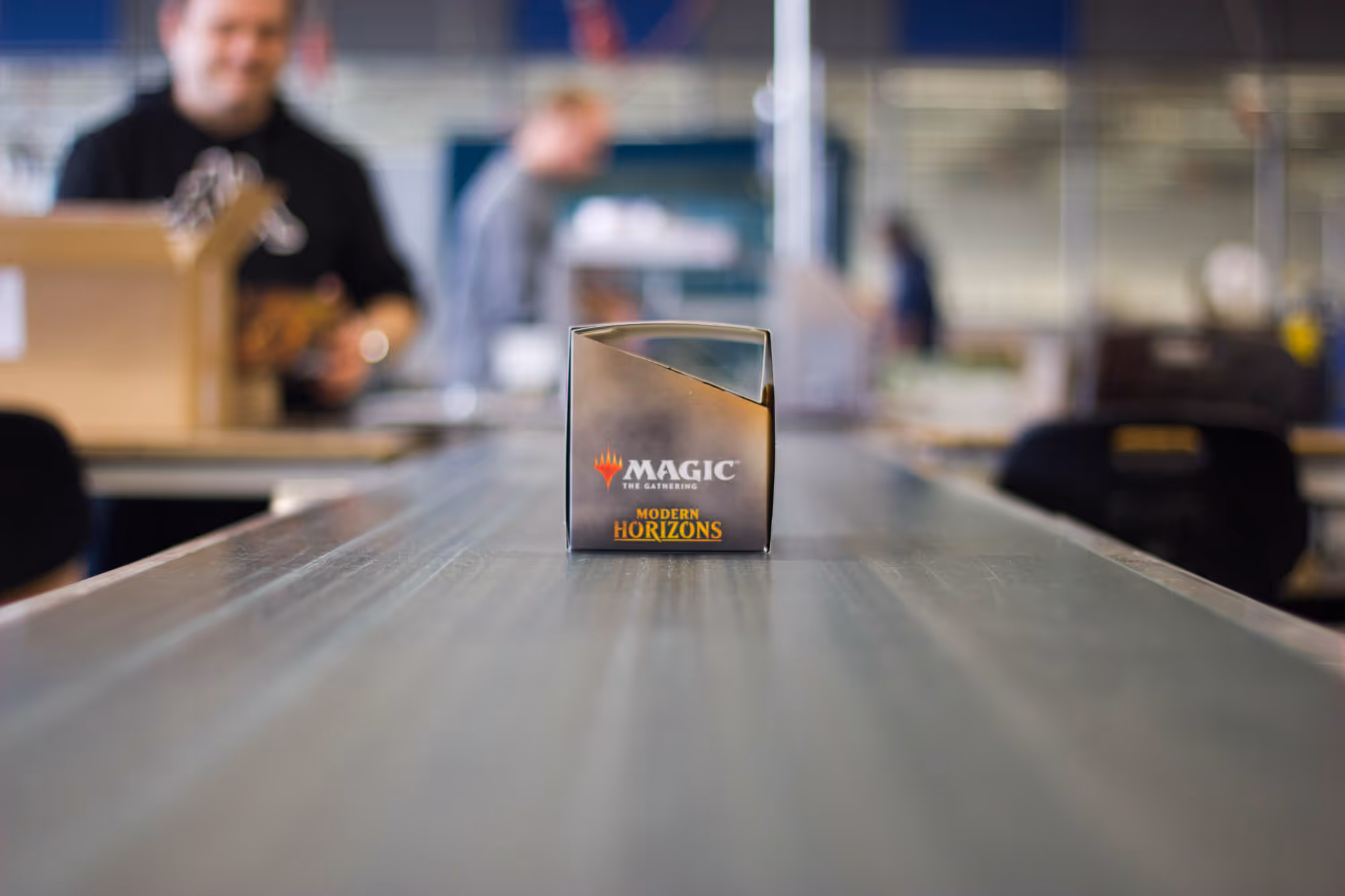 A box of Magic: The Gathering "Modern Horizons" takes center stage on a long table in the middle. In the background, employees are busy packing boxes in a busy warehouse.