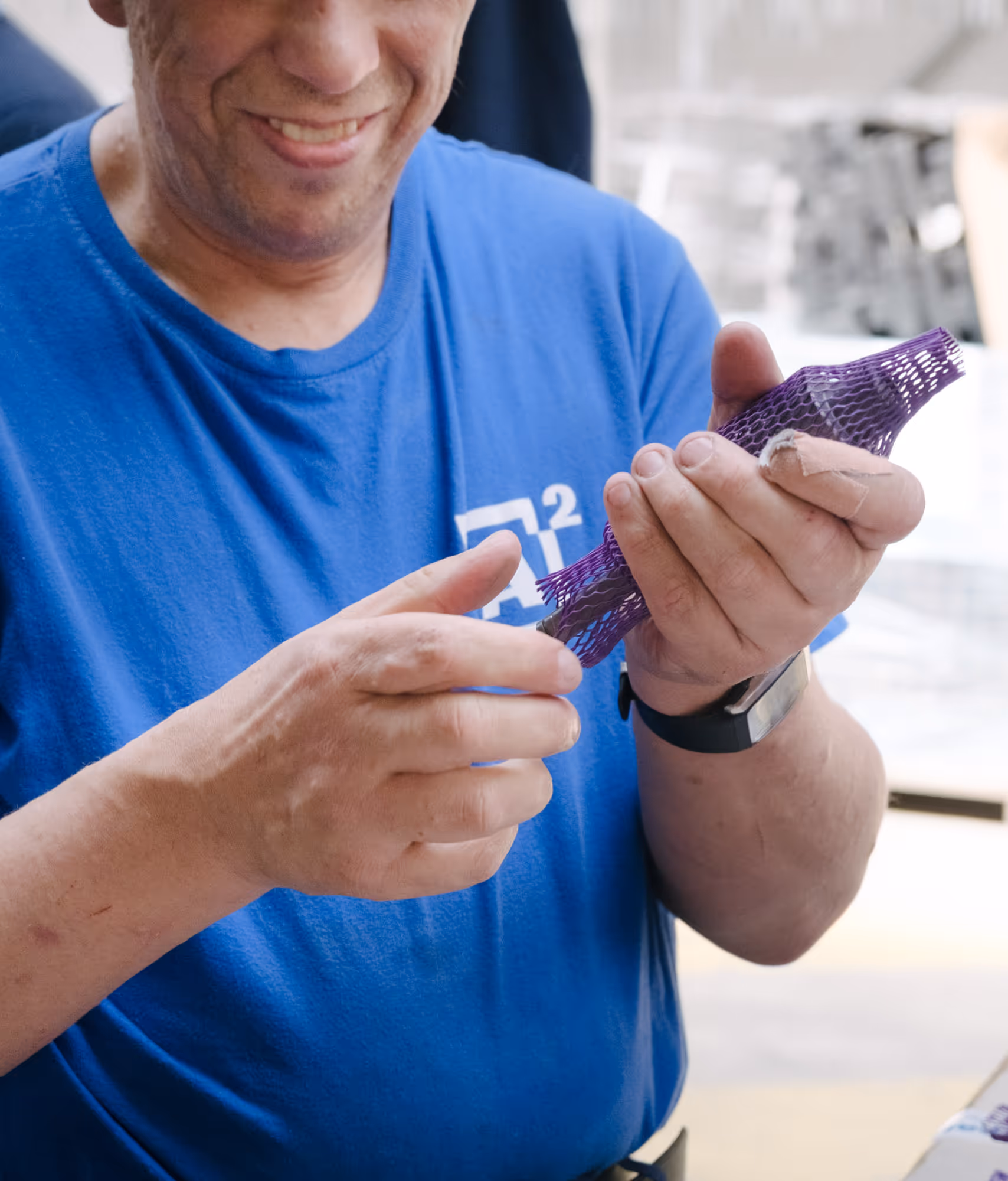 A person in a blue shirt, smiling while holding a piece of purple, gauze-like material, which radiates curiosity and interest. The setting appears informal.