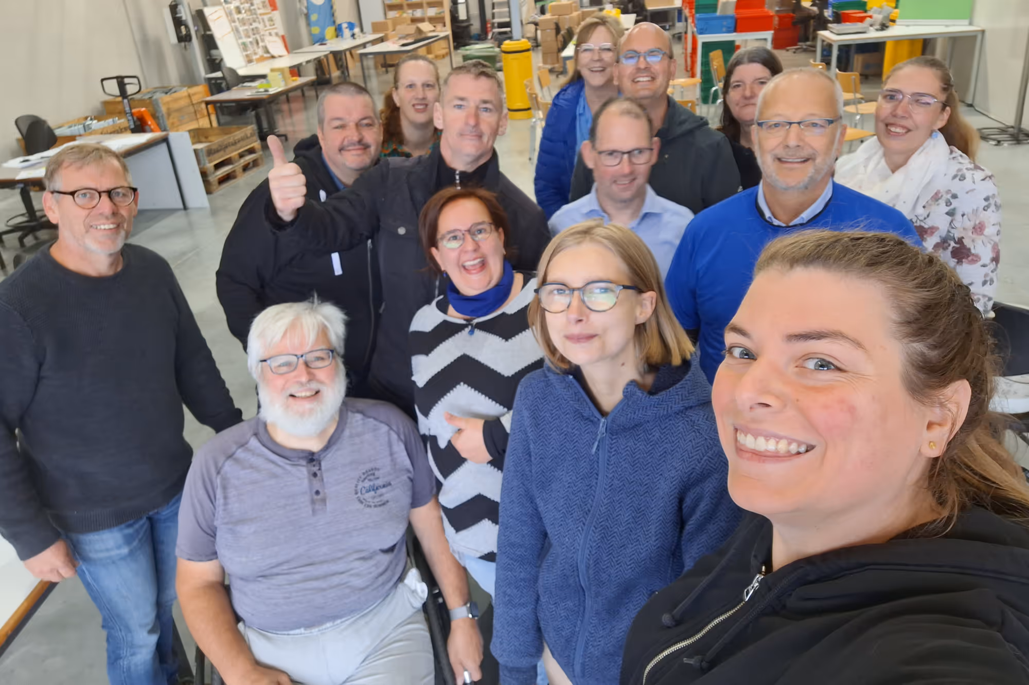 Group of thirteen adults smiling in a brightly lit workshop, with one person giving a thumbs up. The atmosphere is cheerful and collegial.